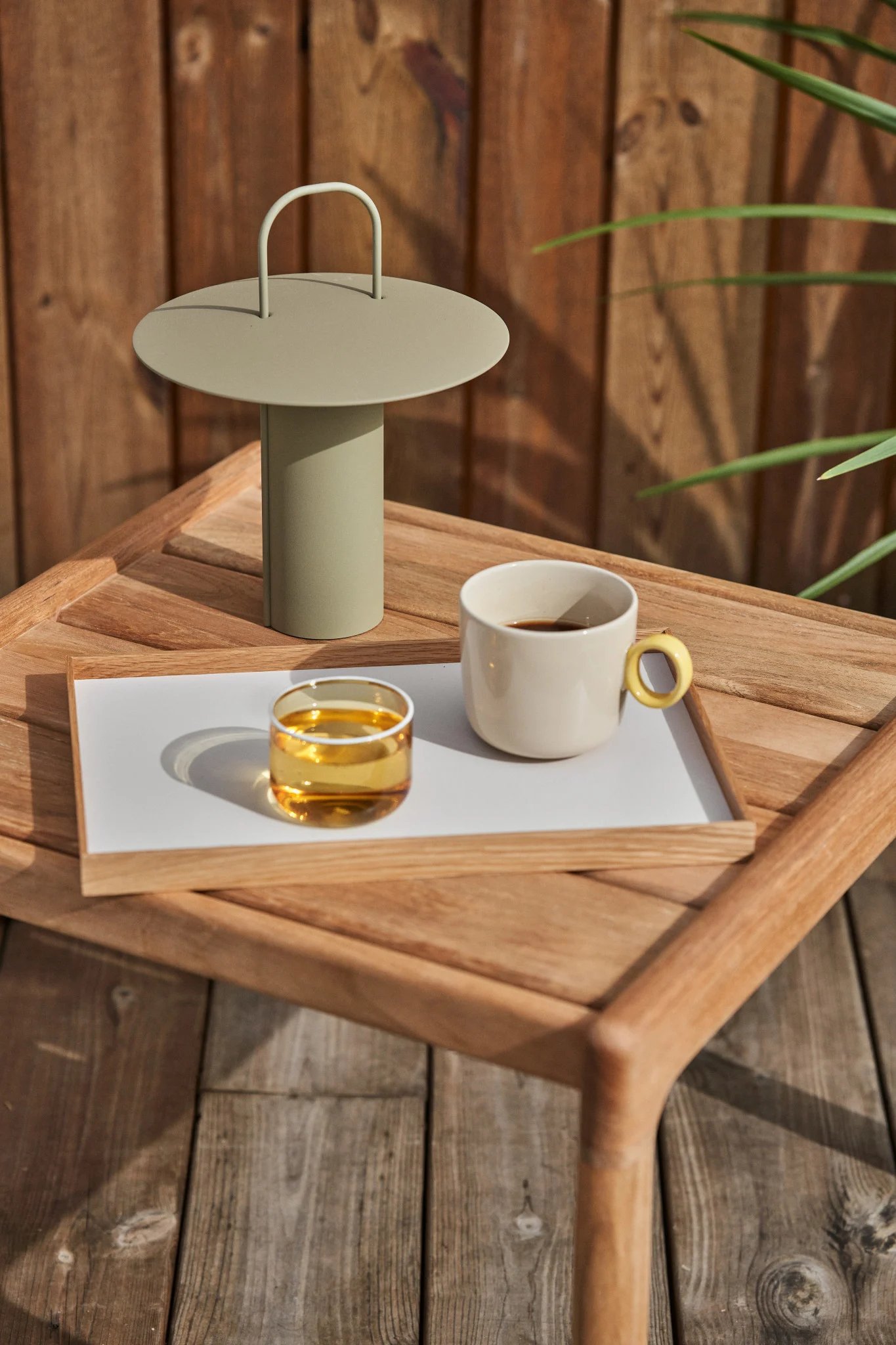 En ljusgrön bärbar lampa, ett glas bärnstensfärgad vätska och en krämfärgad kaffemugg på en träbricka på ett utomhusbord i trä.