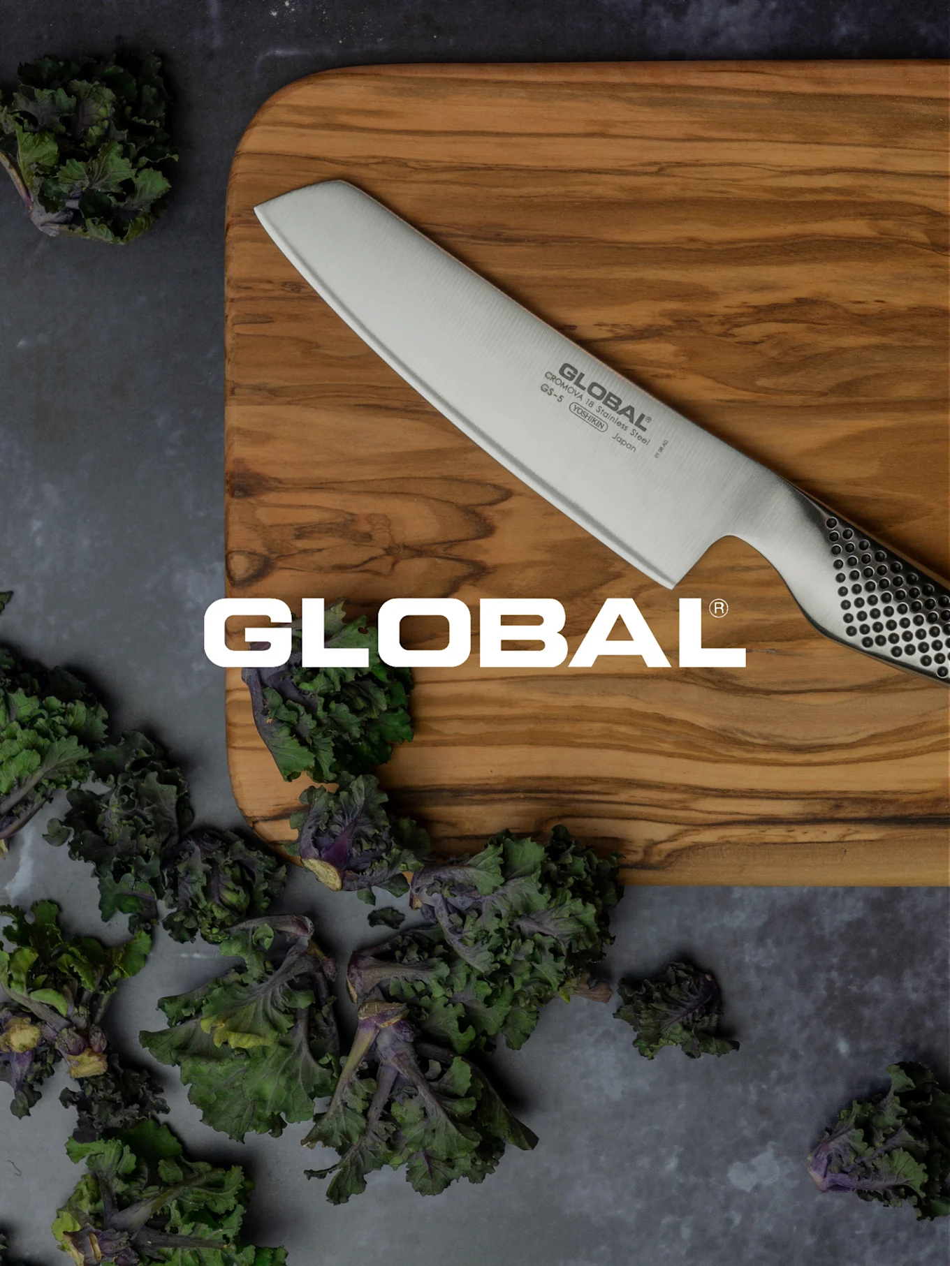 En Global-kniv på en skärbräda i trä, omgiven av färska gröna och lila bladgrönsaker.