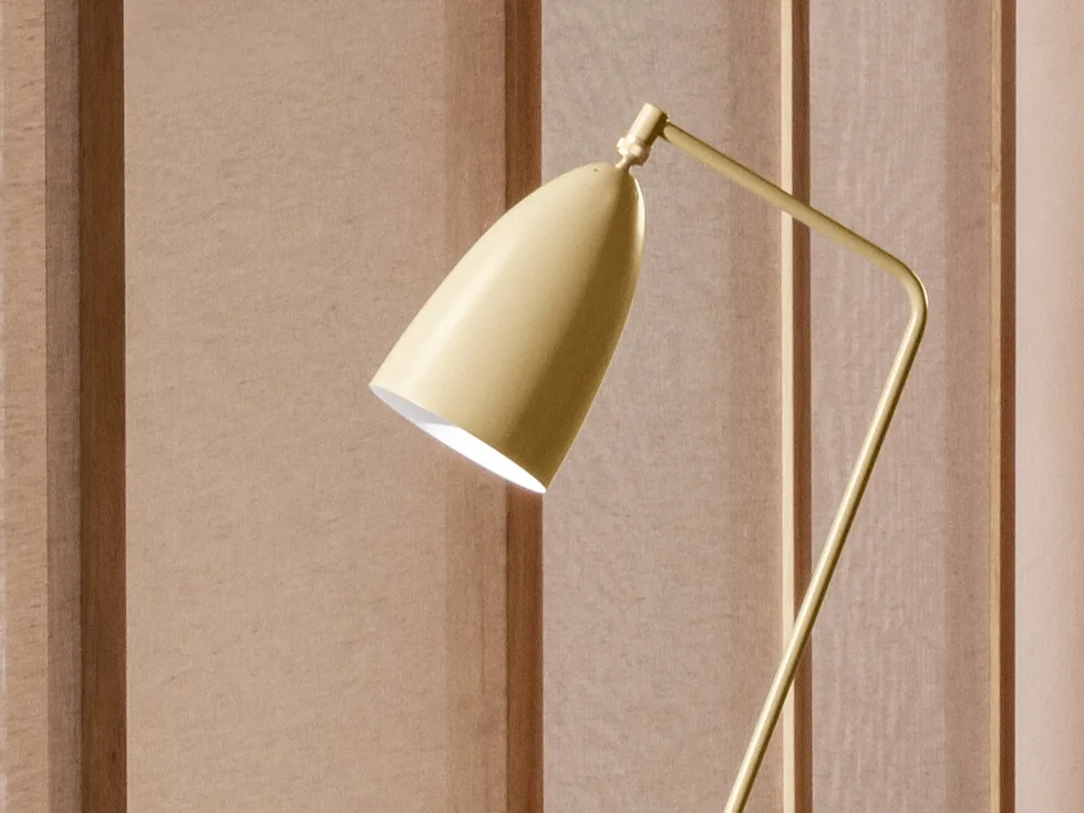 En närbild av en gulaktig-guld modern lampa med ett koniskt huvud vinklat nedåt, som avger ljus mot en ljusbrun panelvägg.