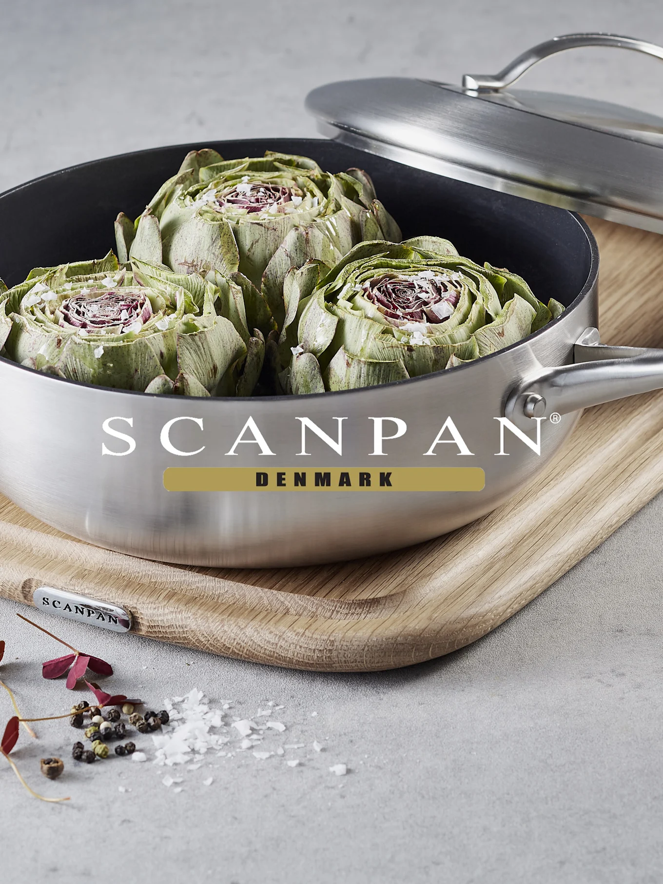 Tre halverade kronärtskockor med salt i en borstad rostfri kastrull på en träskärbräda. Spridda pepparkorn och saltflingor i närheten.