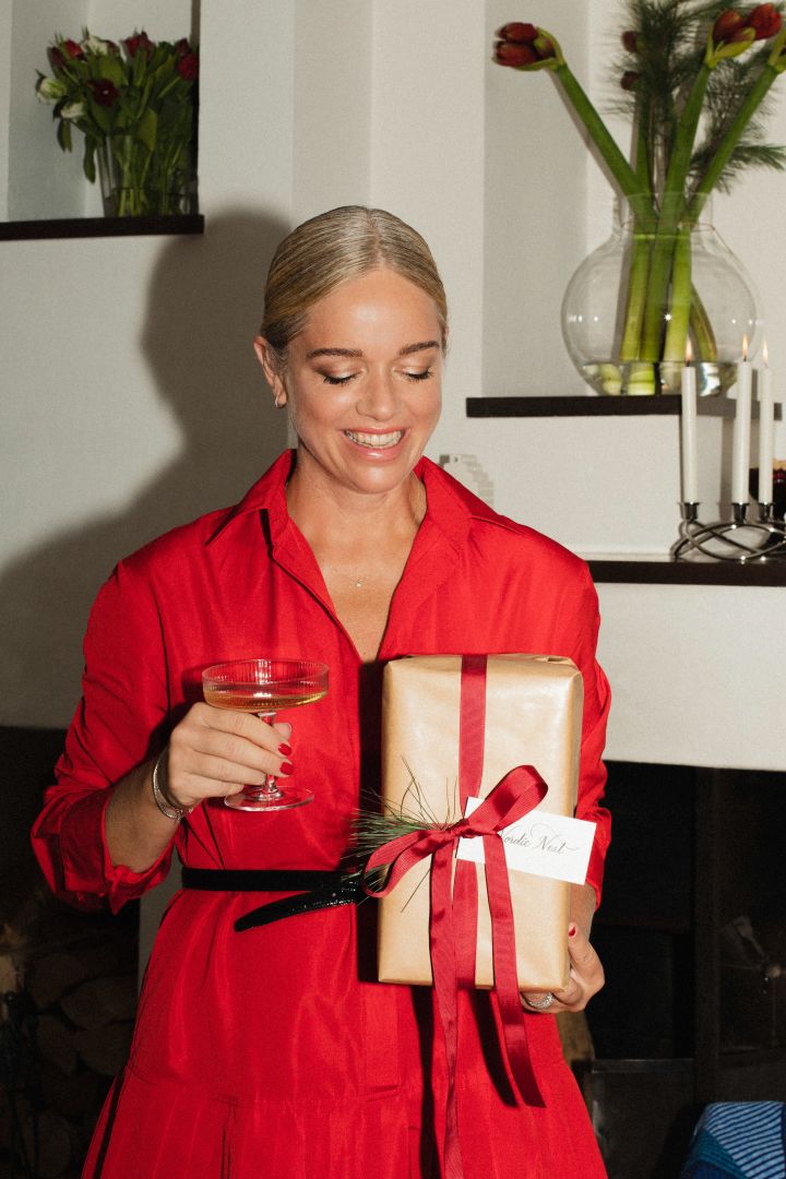 Ebba Kleberg von Sydow håller i ett paket och ett Ripple champagneglas från ferm LIVING i handen klädd i en vacker klänning i rött.