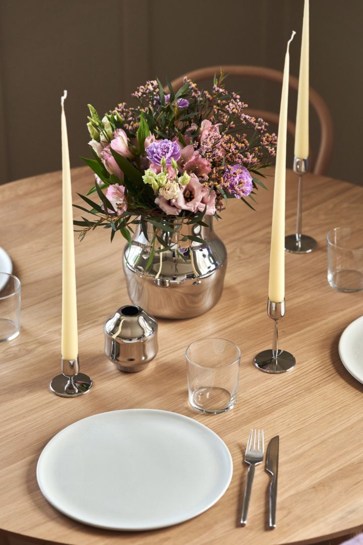 En spegelblank vas i rostfritt stål står mitt på ett bord med en bukett färska lila blommor. 