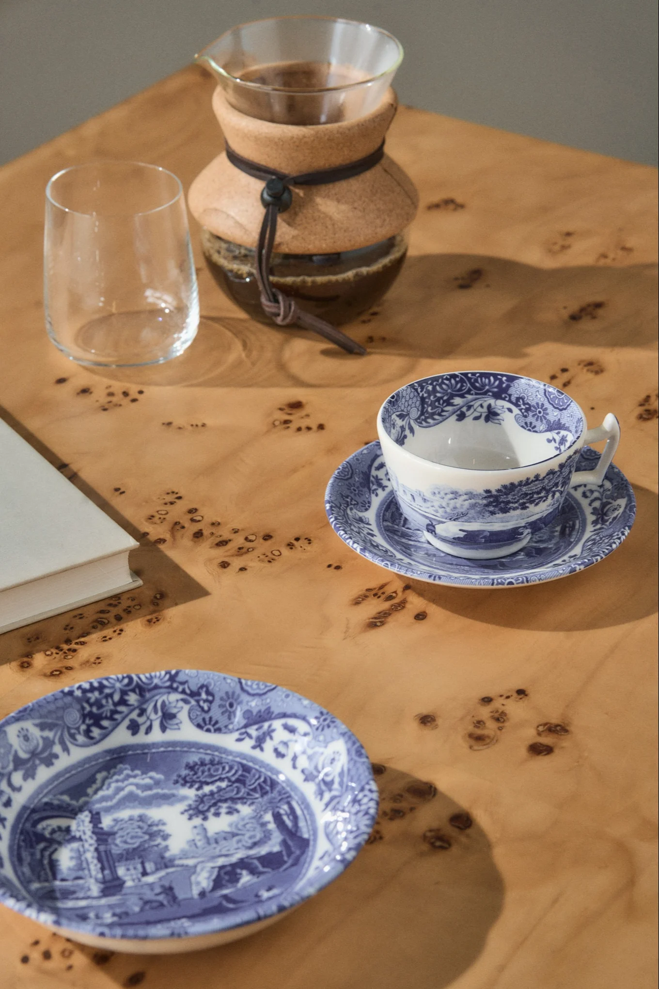 En pour-over kaffebryggare, ett tomt glas, en blå- och vitmönstrad tekopp på ett fat, och en matchande tallrik på ett ljust träbord.