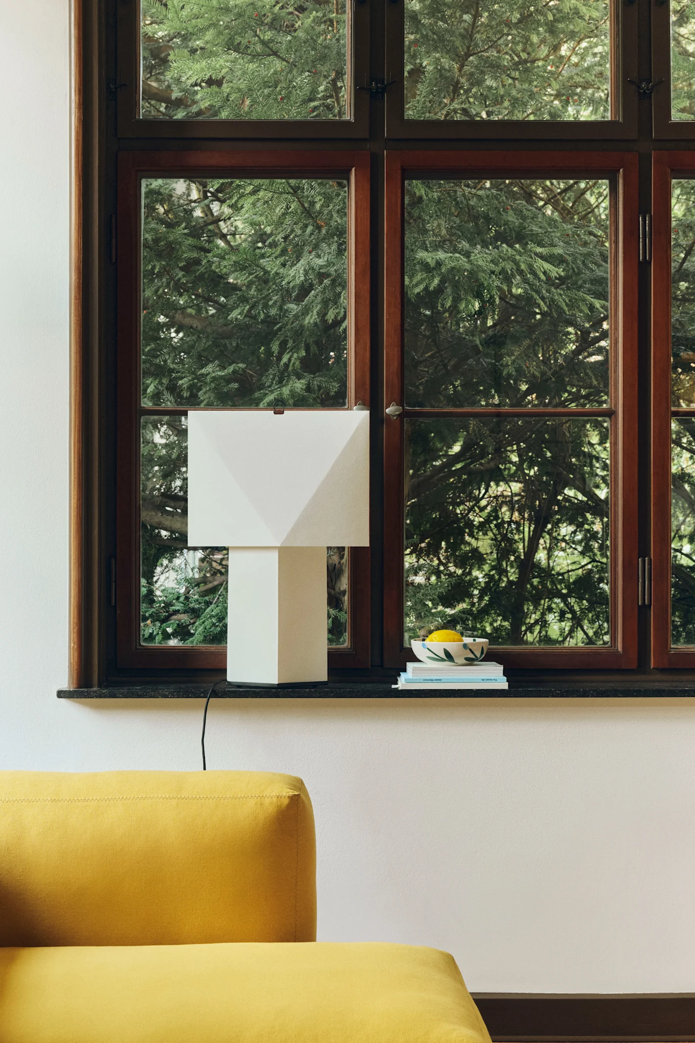 En geometrisk vit bordslampa på en mörk fönsterbräda bredvid en skål med citroner och böcker, med en gul soffa i förgrunden.