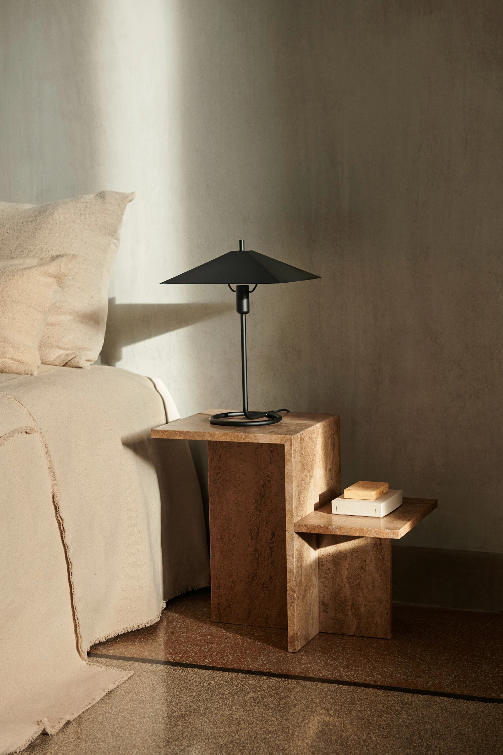 En sovrumsscen med en beige säng bredvid ett texturerat brunt sidobord i två nivåer, toppat med en svart modern bordslampa.