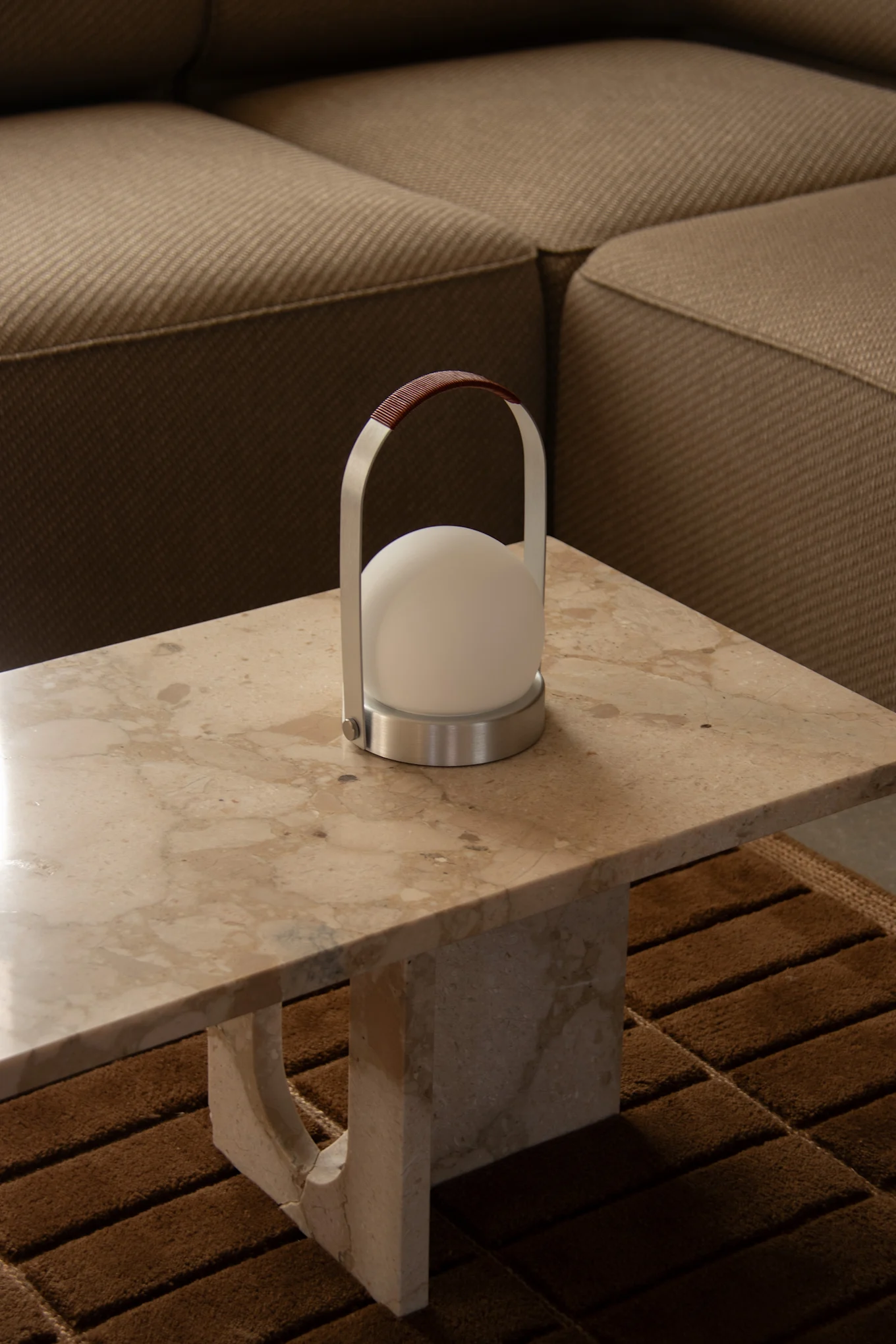En modern bärbar lampa med en vit sfärisk lampa, borstad metallram och brunt läderhandtag står på ett ljust marmorbord.