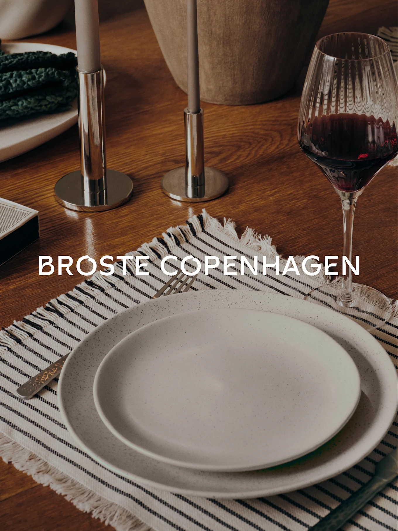 Matbord med två spräckliga tallrikar på en randig bordstablett, gaffel, vinglas, två ljusstakar och texten 'BROSTE COPENHAGEN'.
