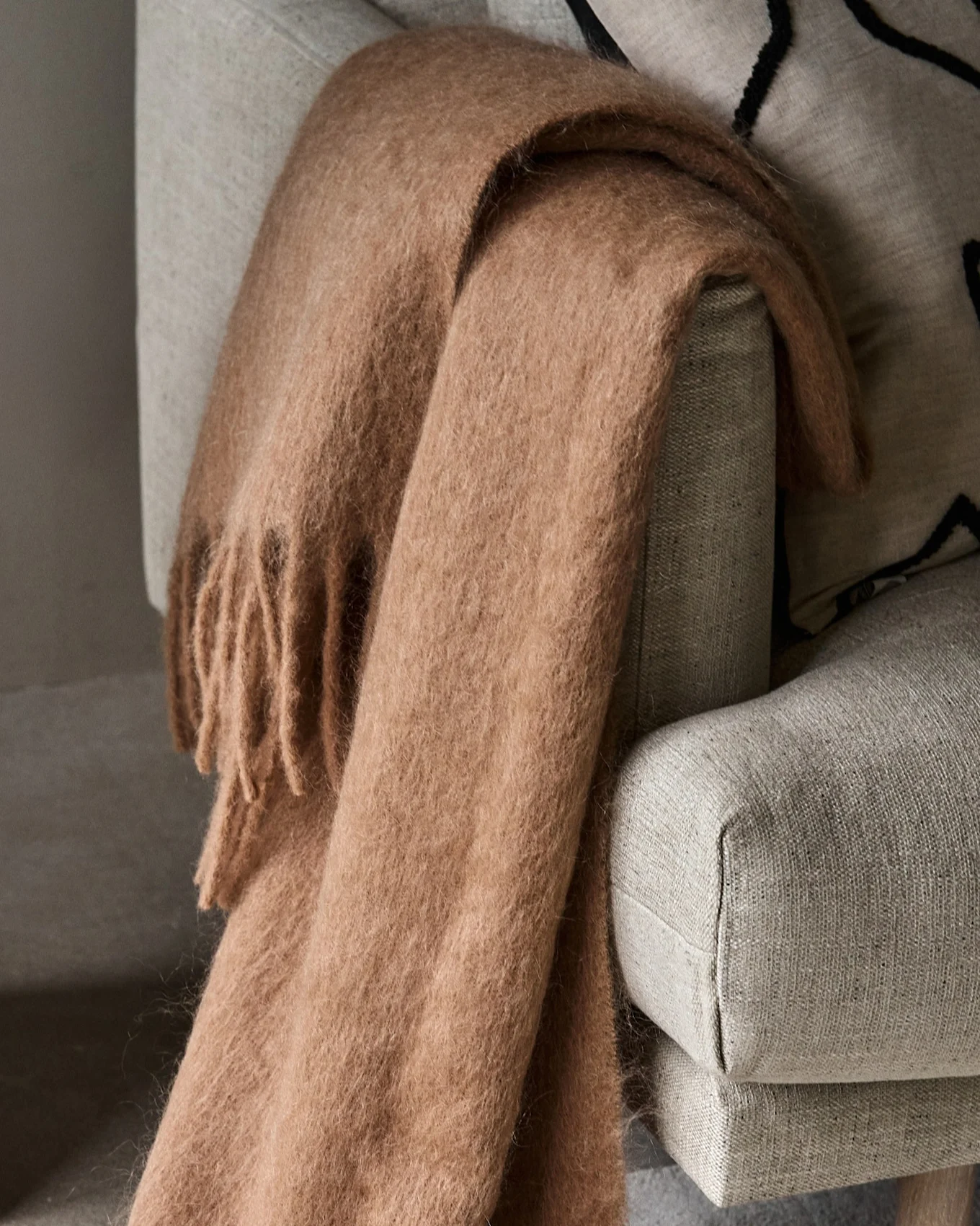 Mysig ljusbrun fluffig pläd med fransar draperad över en ljusgrå fåtölj.