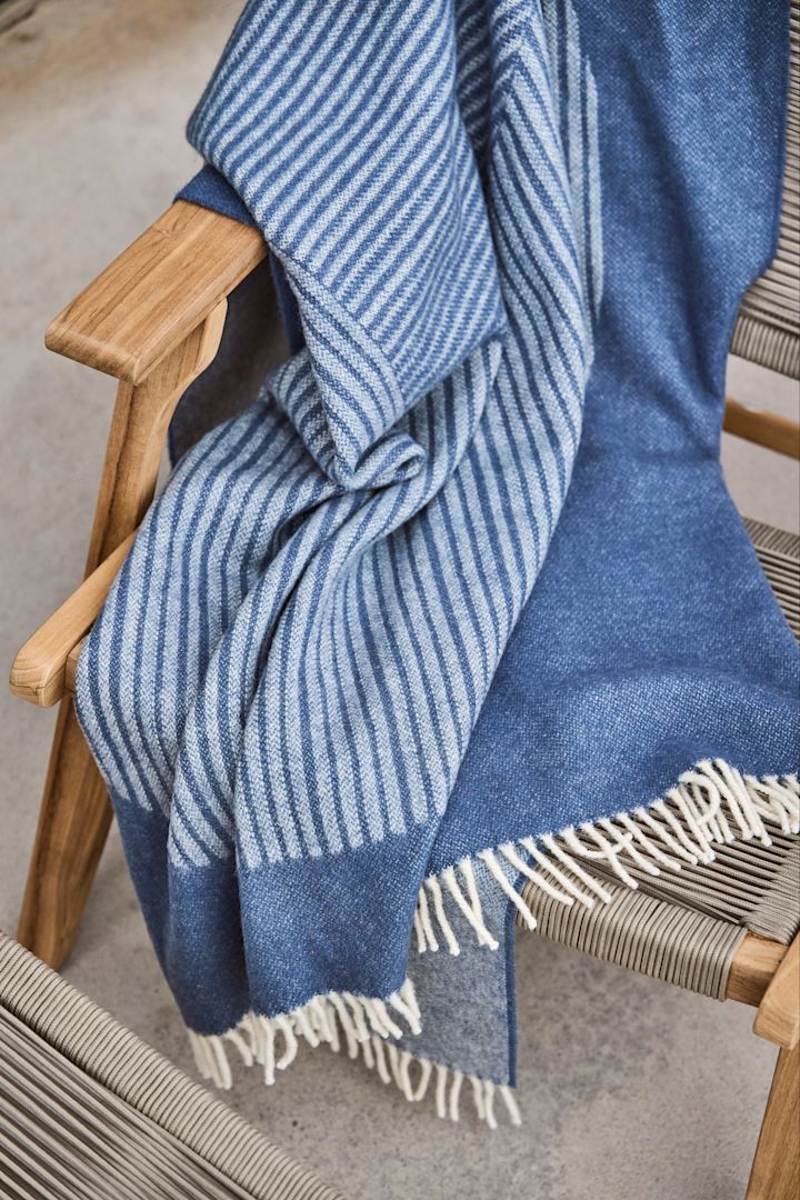 Stripes ullpläd från NJRD i en blå färg med randigt mönster ligger över en stol i trä.