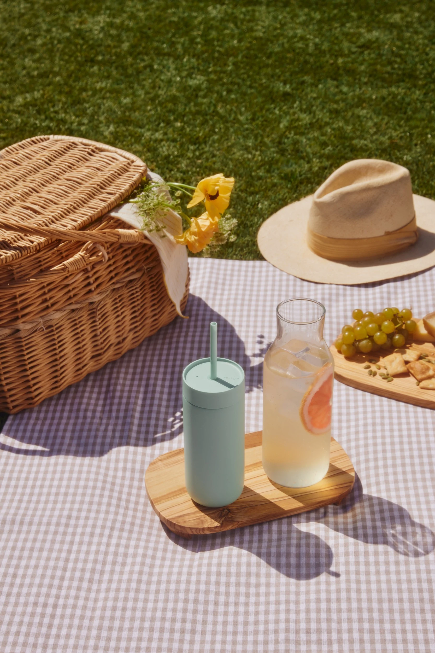 Picknick på rutig filt: korg, ljusgrön mugg, grapefruktdryck, vindruvor och kex på gräset.