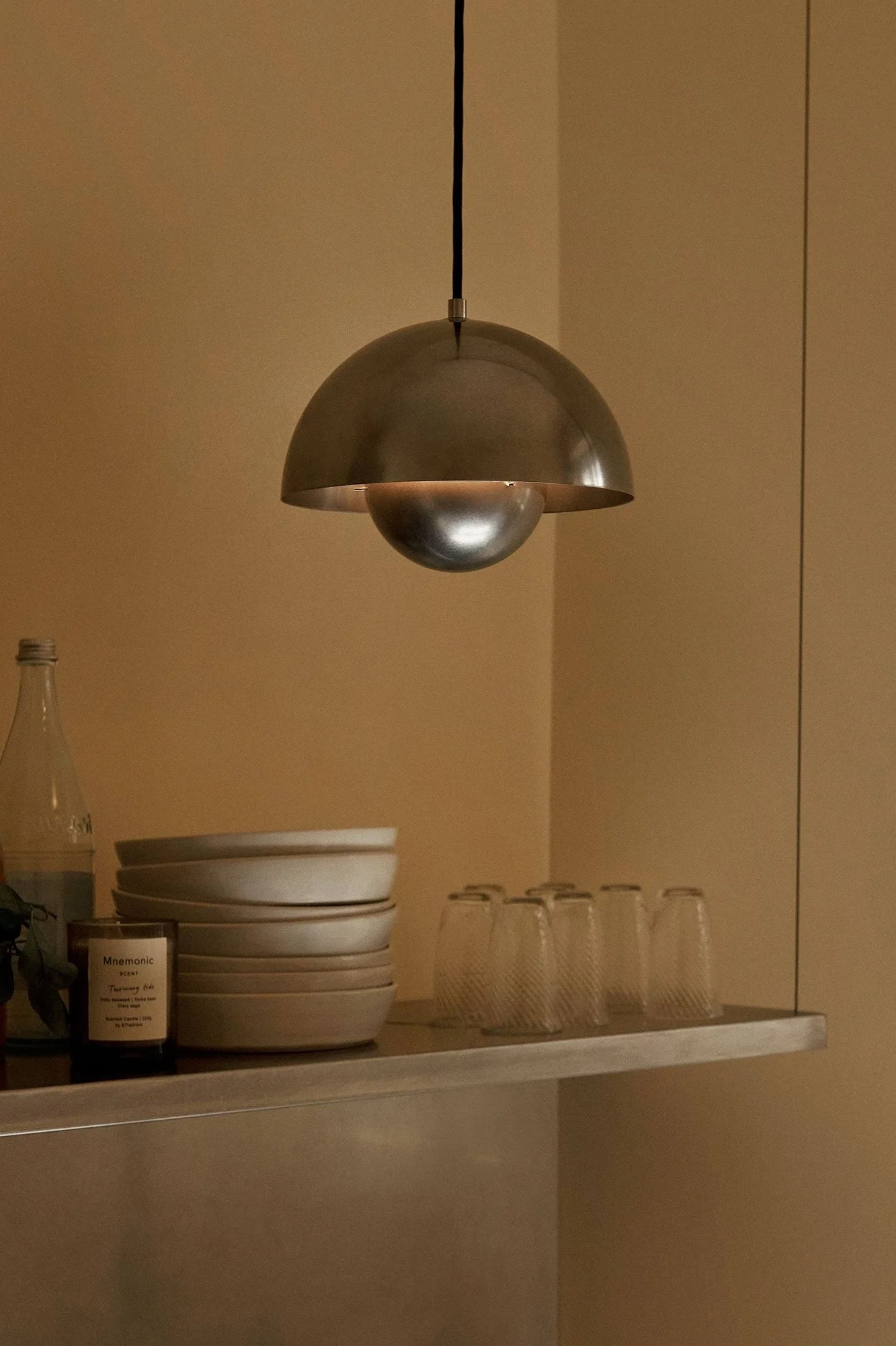 Metallisk kupolpendellampa lyser upp en hylla med staplade skålar, glas, en flaska och ett ljus i varmt rum.