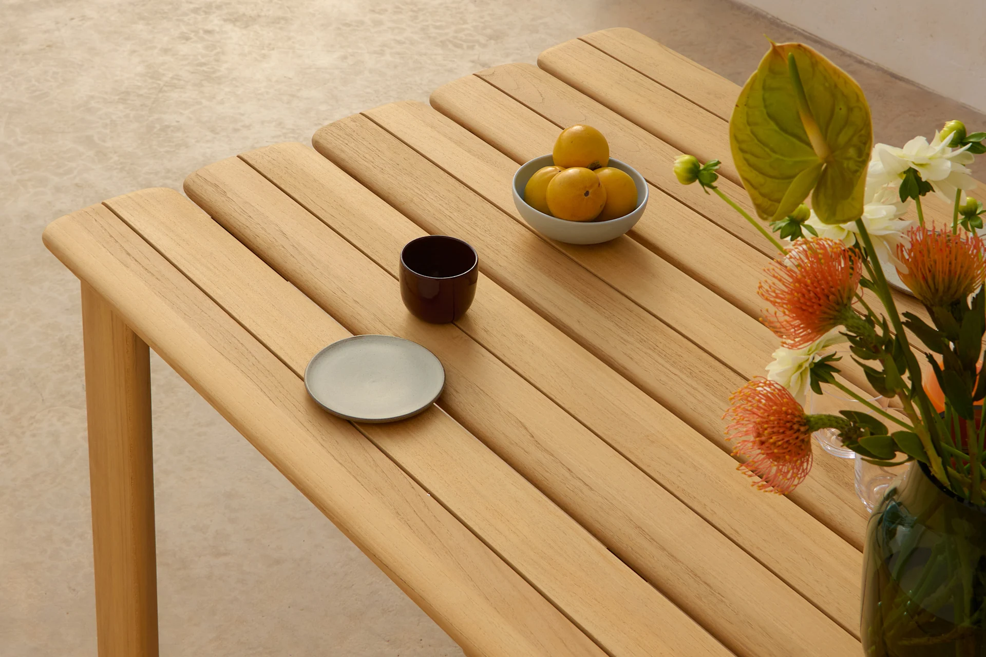 Ett ljust träbord med ribbor, med en skål gula frukter, en mörk kopp, en grå tallrik och en vas med orange och vita blommor.