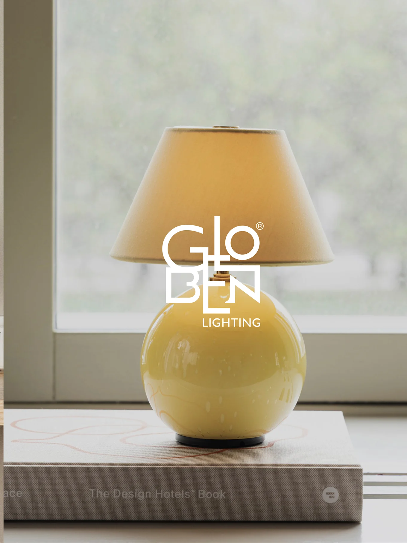 Gul bordslampa med beige skärm, står på böcker vid ett fönster.