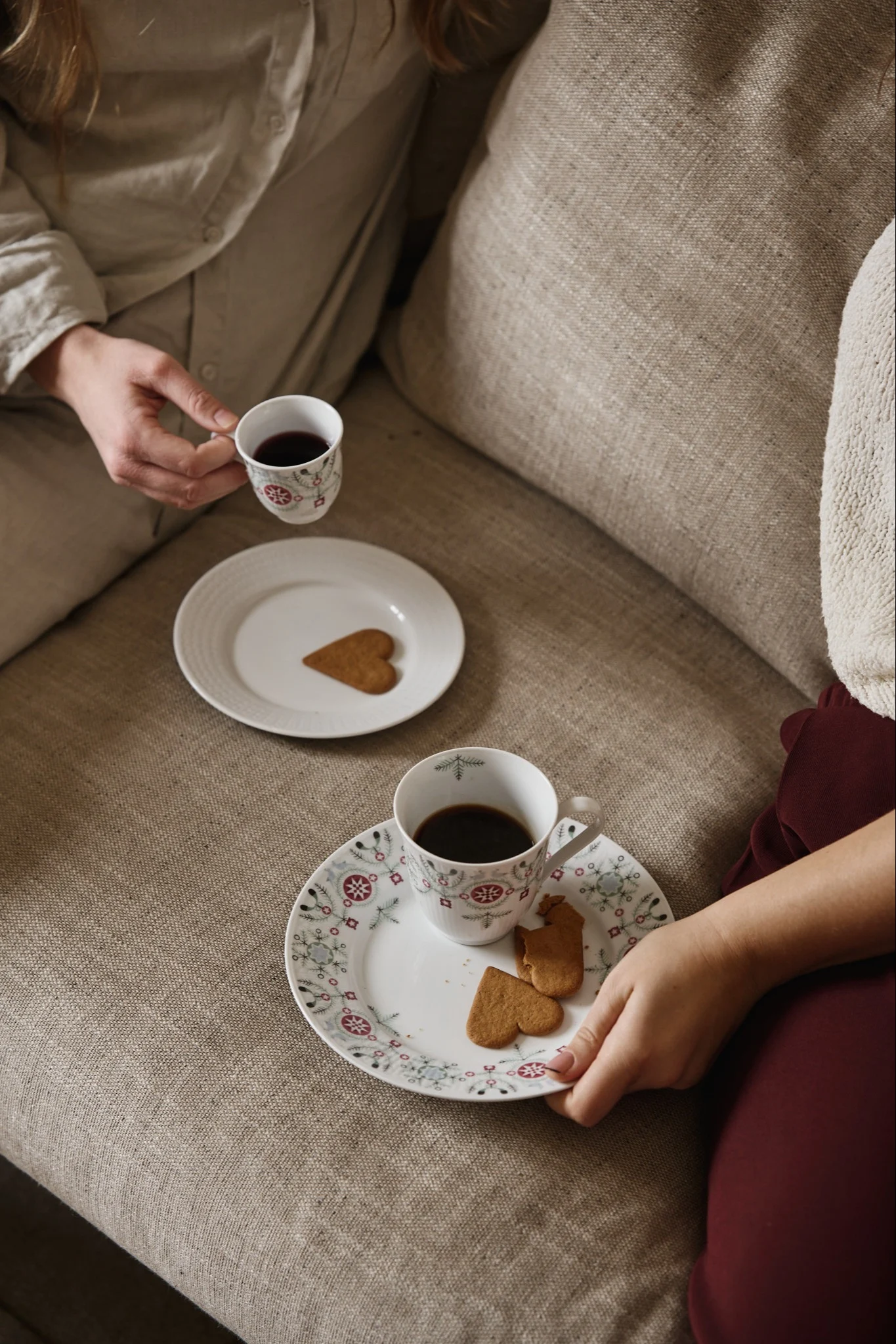 Ett par personer sitter i en soffa med Swedish Grace Winter-muggkollektion. Här ser du både en vanlig mugg och en glöggmugg samt tallrikar med pepparkakor. 