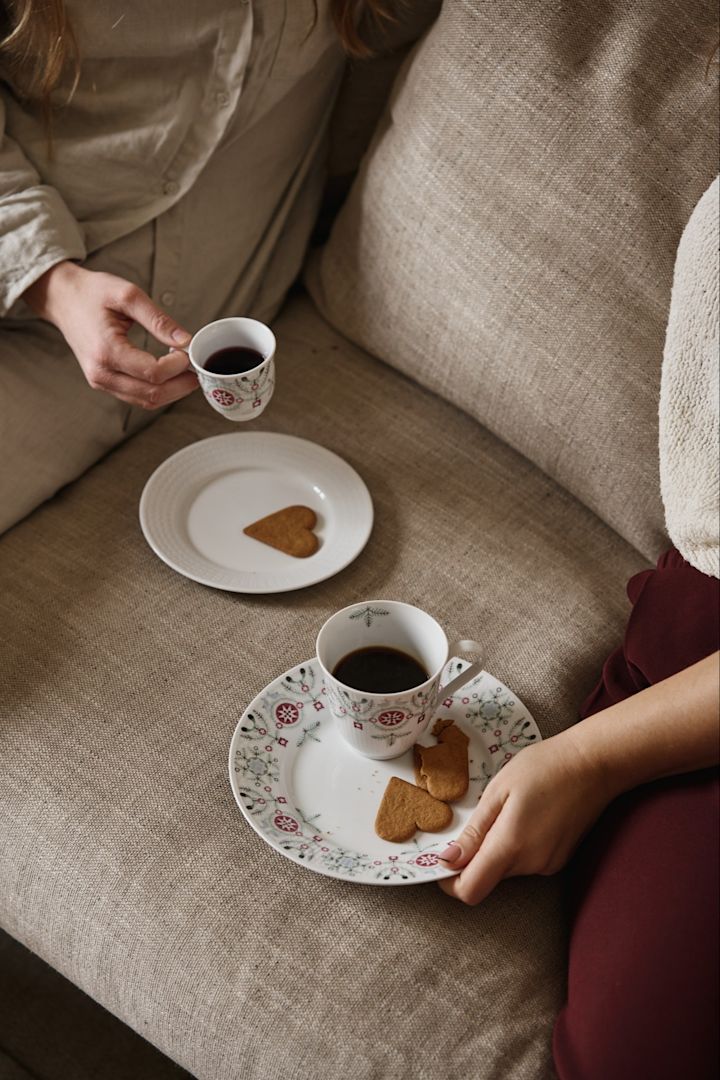 Ett par personer sitter i en soffa med Swedish Grace Winter-muggkollektion. Här ser du både en vanlig mugg och en glöggmugg samt tallrikar med pepparkakor. 