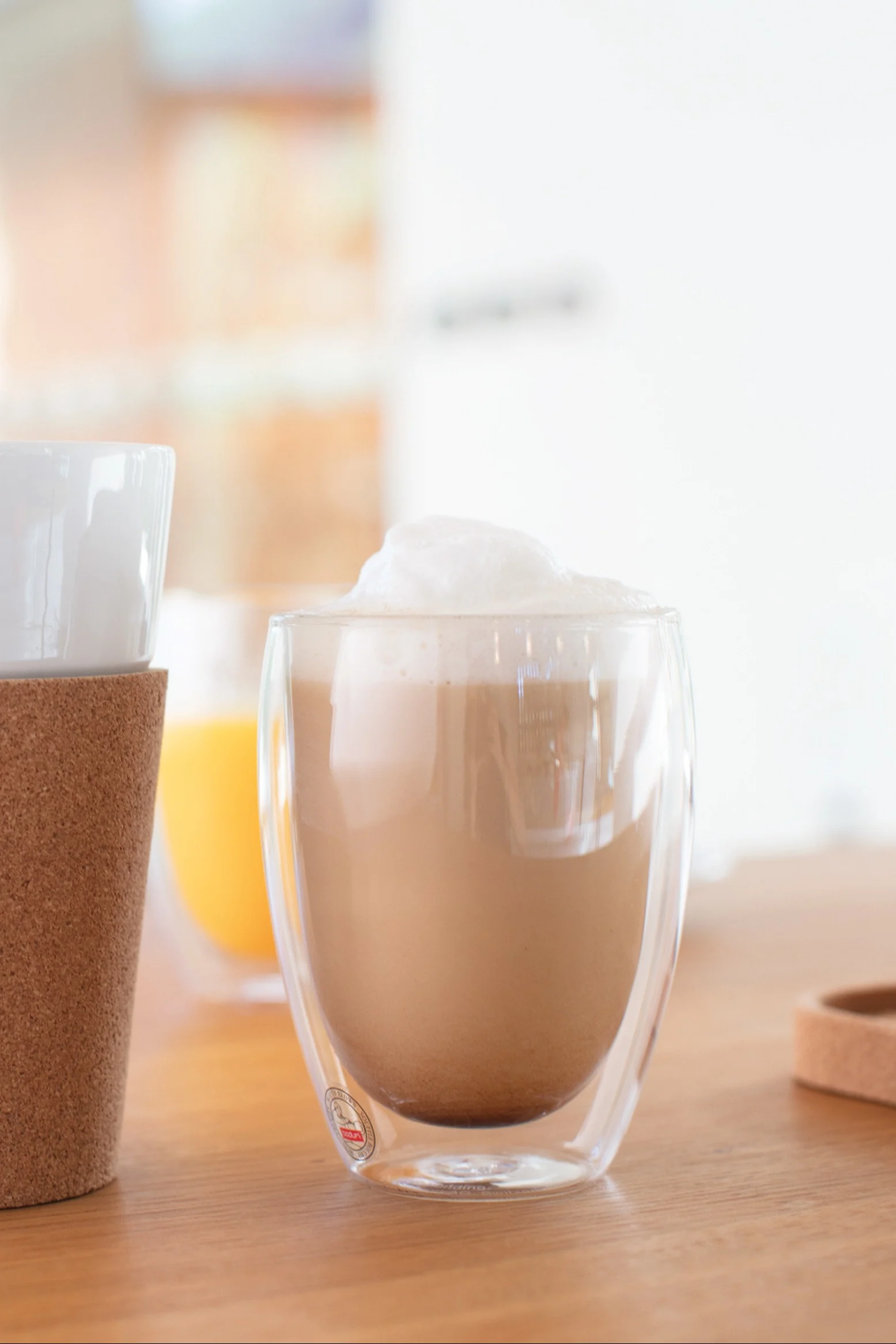 Skummig latte i en dubbelväggig klar glasmugg på ett träbord, med en annan mugg med korkärm till vänster och en apelsindryck i bakgrunden.