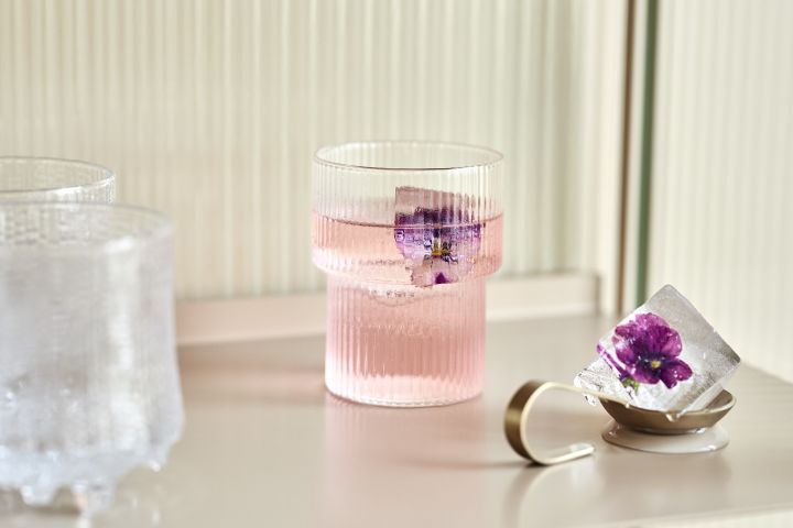 Ripple dricksglas från Ferm Living med en rosa drink i och lila blomma fryst i en isbit. 