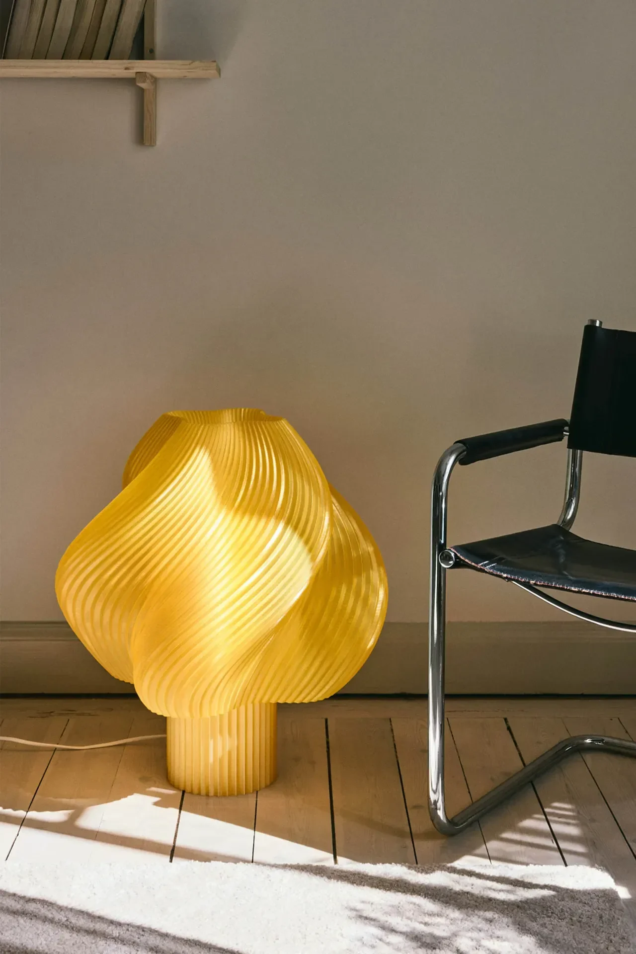 En ljust gul, skulptural bordslampa med räfflad, vågig design lyser på ett trägolv bredvid en svart läder- och kromstol.