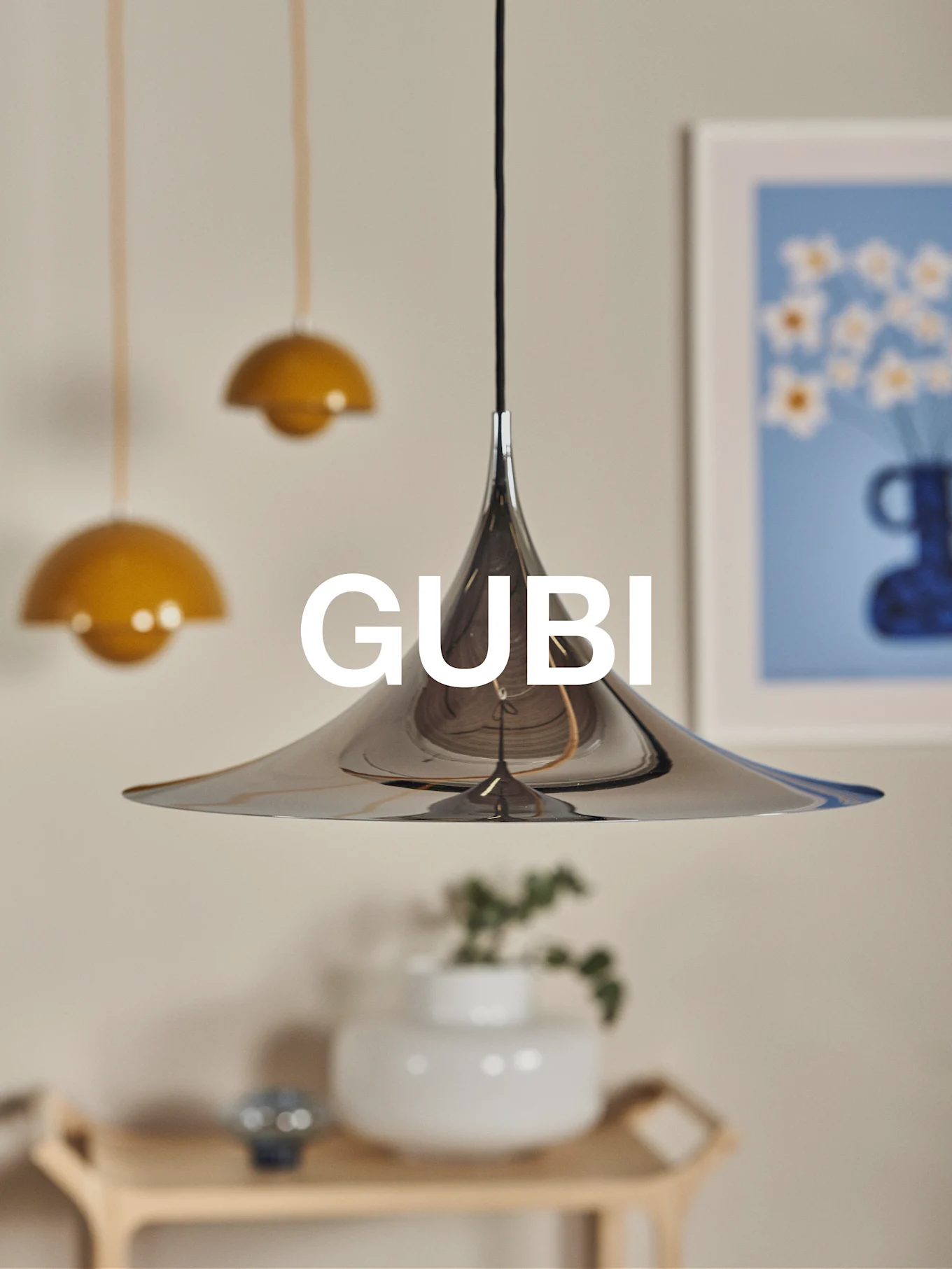 En polerad kromkonisk taklampa med GUBI-logotyp, mot en suddig bakgrund med andra lampor och konst.