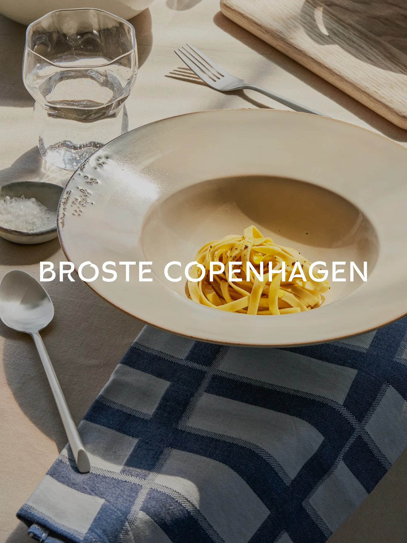 Pasta i en bred keramikskål, med havssalt, ett texturerat vattenglas, gaffel och sked på en blåmönstrad servett.
