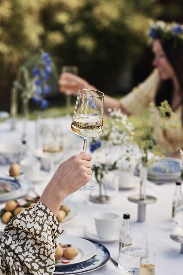 Ett Iittala Essence vitvinsglas hålls upp över ett midsommarbord.
