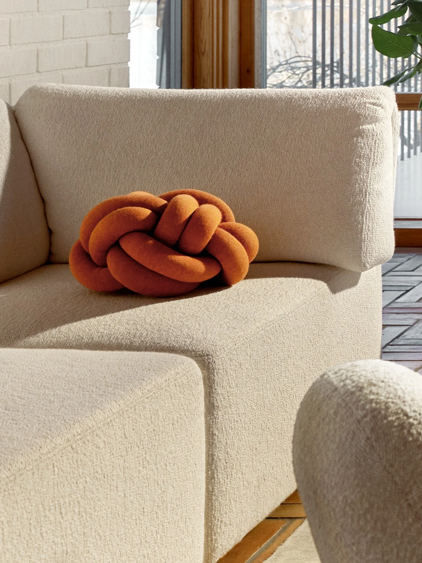En orange knutkudde på en beige texturerad soffa, med en vit tegelvägg och ett fönster i bakgrunden.