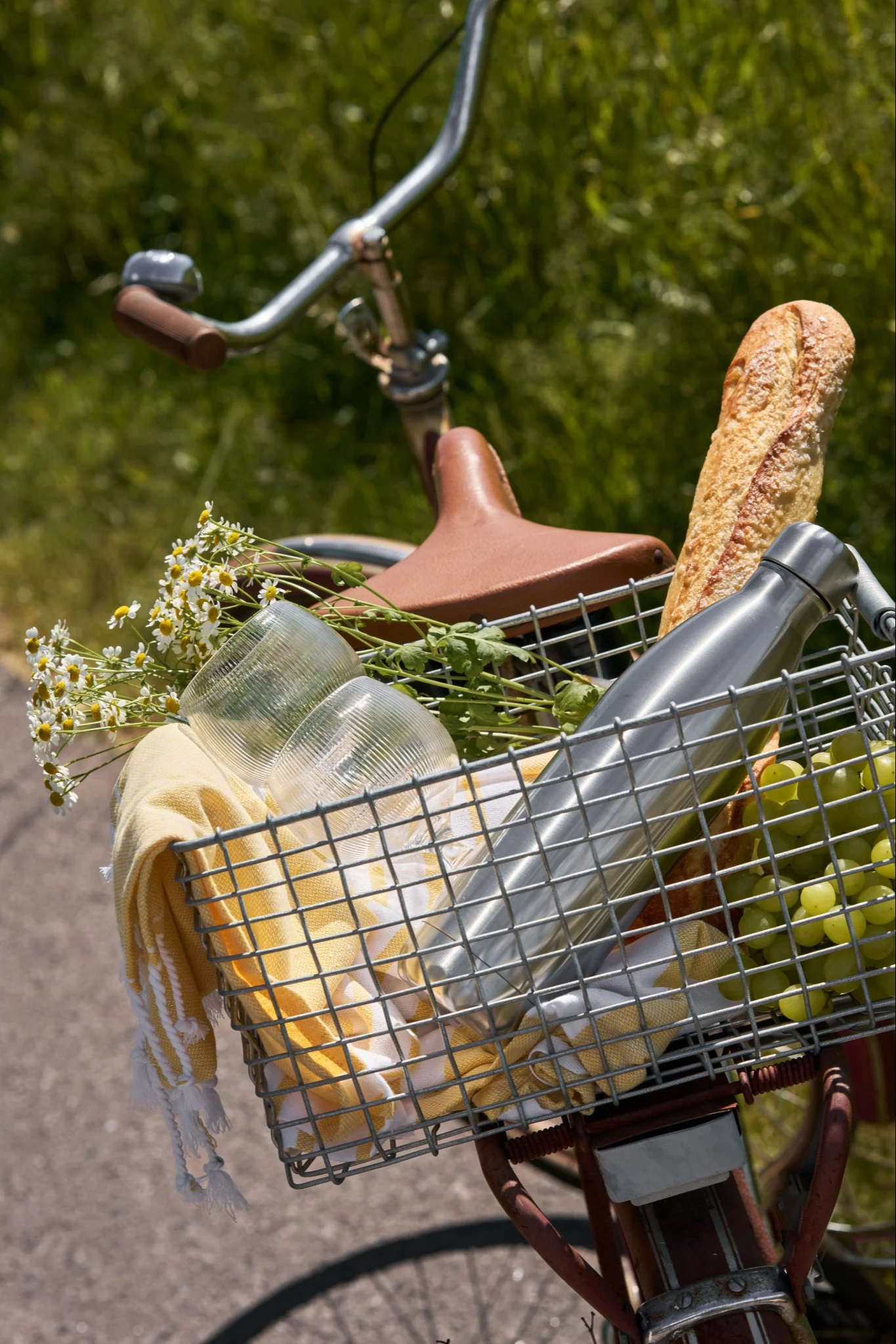 Cykel med en framkorg fylld med picknickartiklar: bröd, gröna druvor, en silverfärgad vattenflaska, muggar, en gul handduk och blommor.
