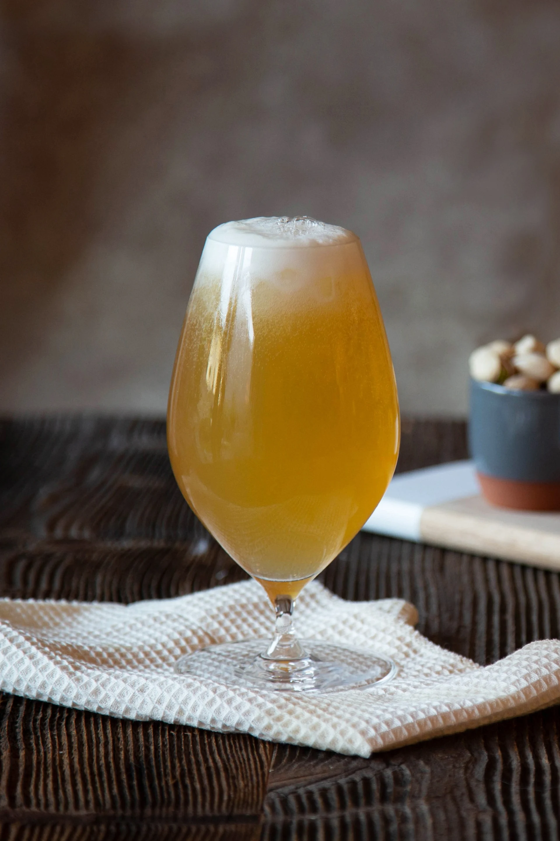 Ett glas skummande bärnstensfärgat öl på en vit duk, med skålar med nötter och brödpinnar i bakgrunden på en träbräda.