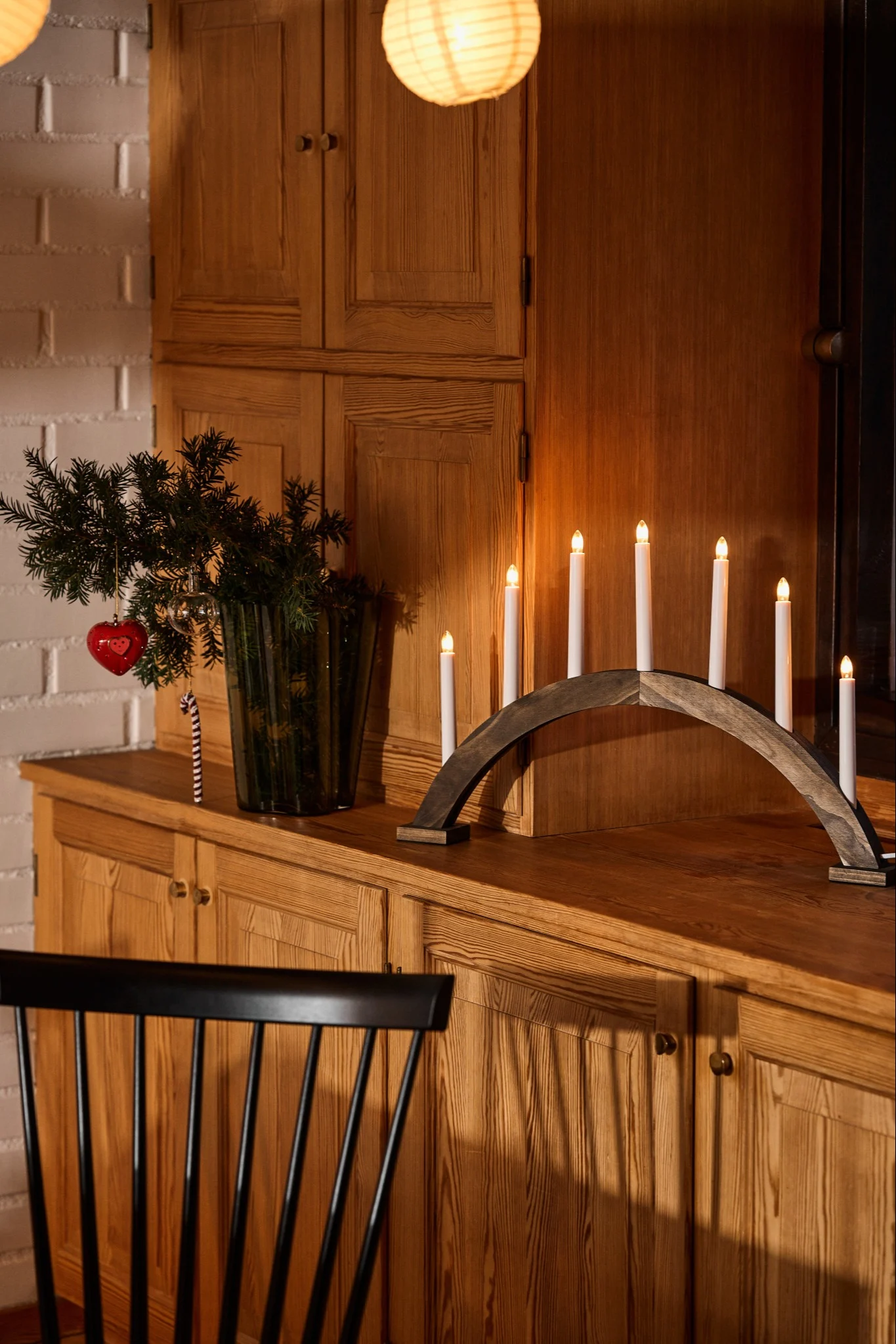 En bågformad adventsljusstake i mörkbetsat trä står på en träbyrå, bredvid en vas fylld med grankvistar.