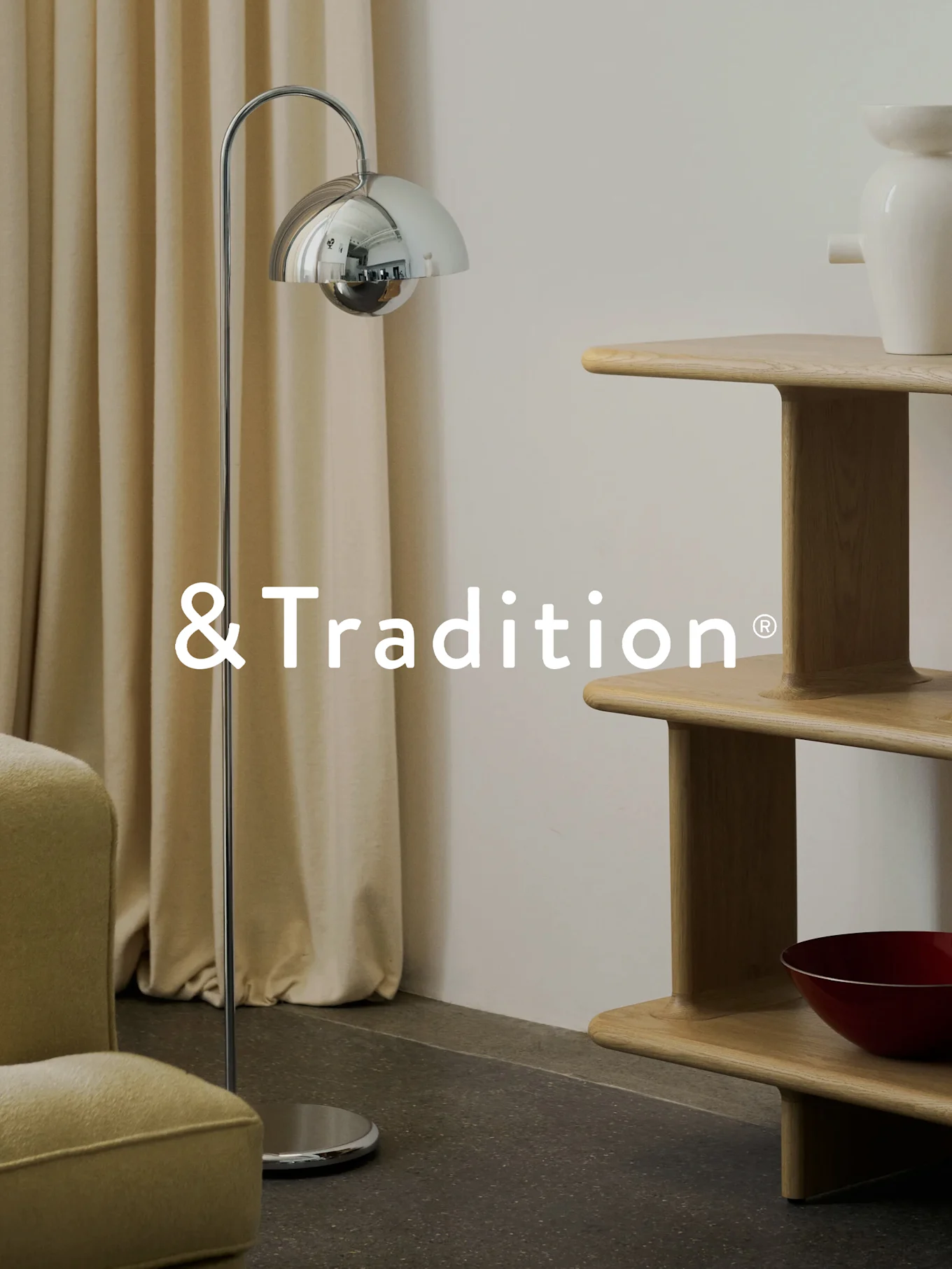 En modern interiör med en kromad golvlampa, en hyllenhet i ljust trä i flera nivåer och en fåtölj. &Tradition® logotyp.