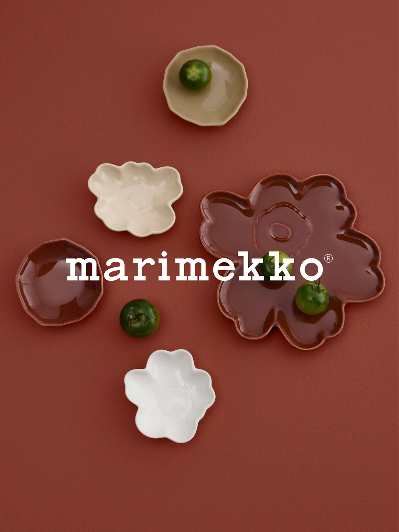 Vit 'marimekko'-logotyp, blomformade fat (vitt, gräddvitt, rödbrunt) med gröna frukter, på rostfärgad yta.