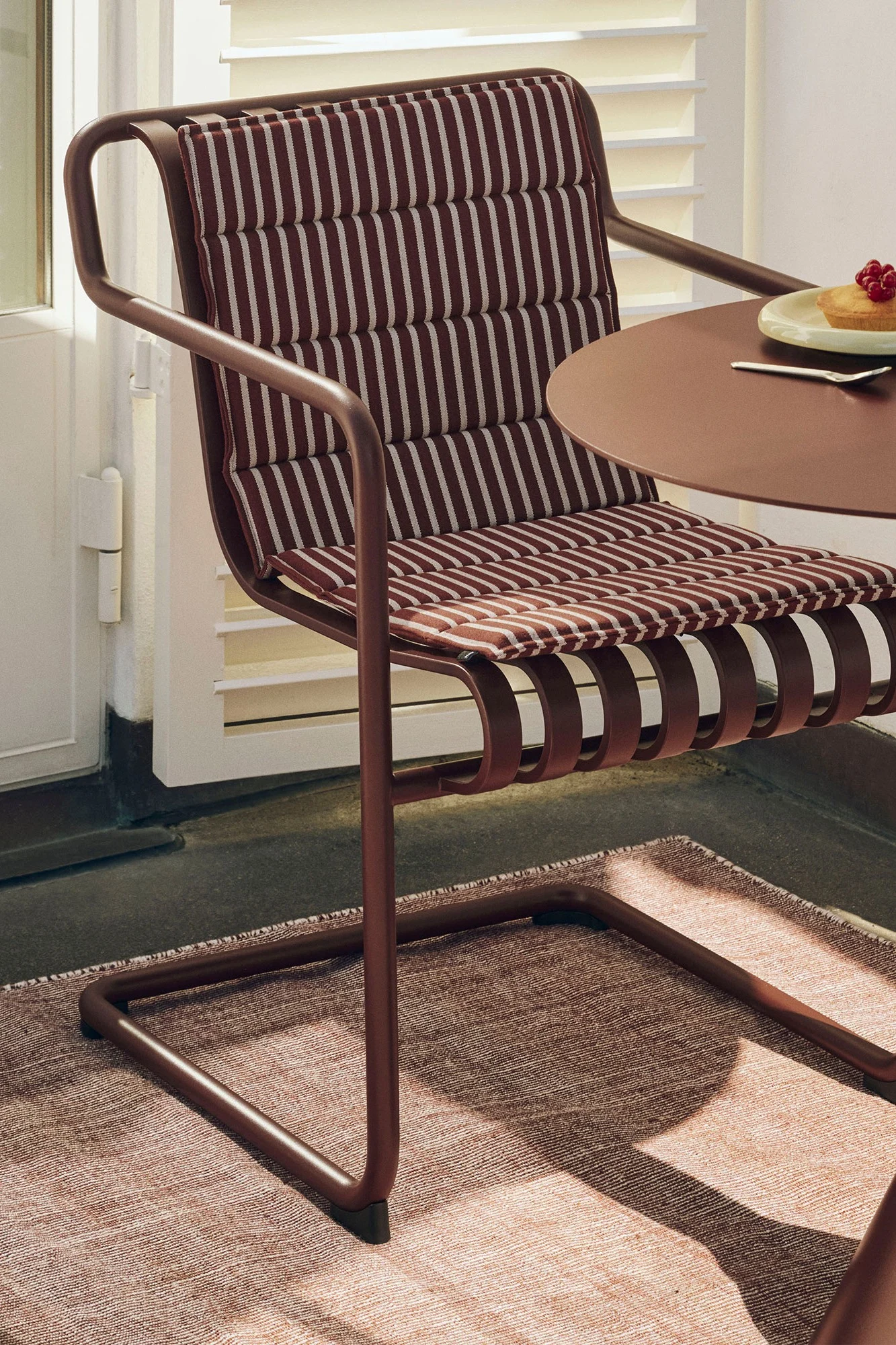 Rödbrun metallstol med randig dyna och matchande bord med tårta på en texturerad matta. Inspiration till din balkong 2026.