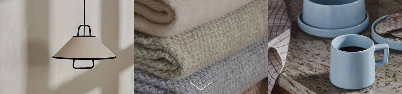 En beige taklampa med svart ram, en hög med filtar i beige, grönt och blått, samt en ljusblå mugg med kaffe på en bänkskiva av granit.