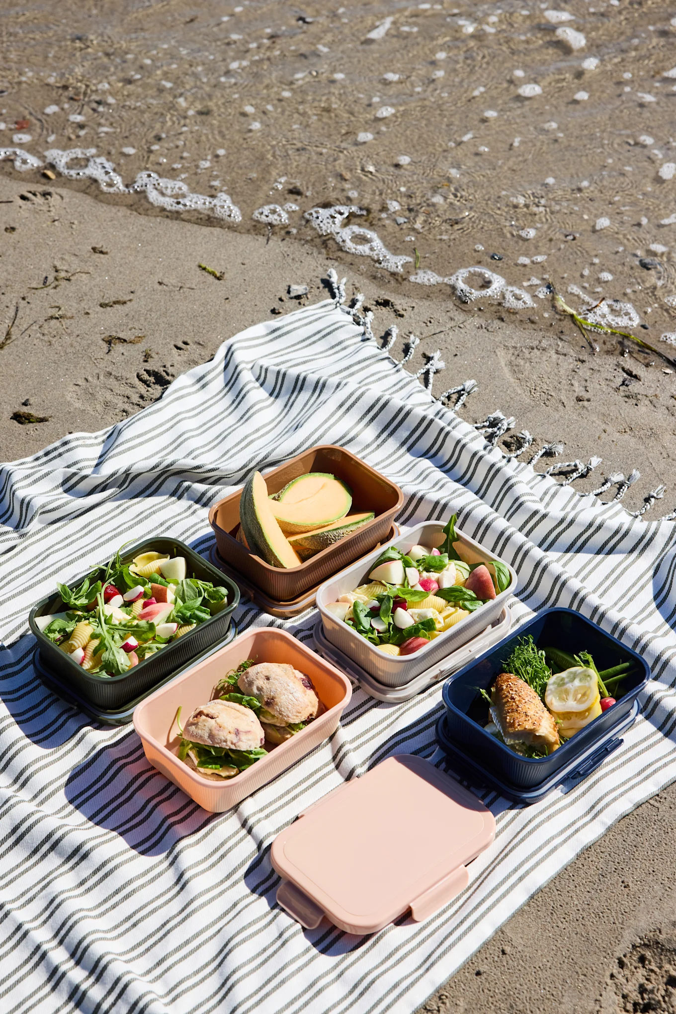 Fem öppna färgglada lunchlådor med sallader, smörgåsar och melon, plus en stängd rosa låda, på en randig strandfilt.