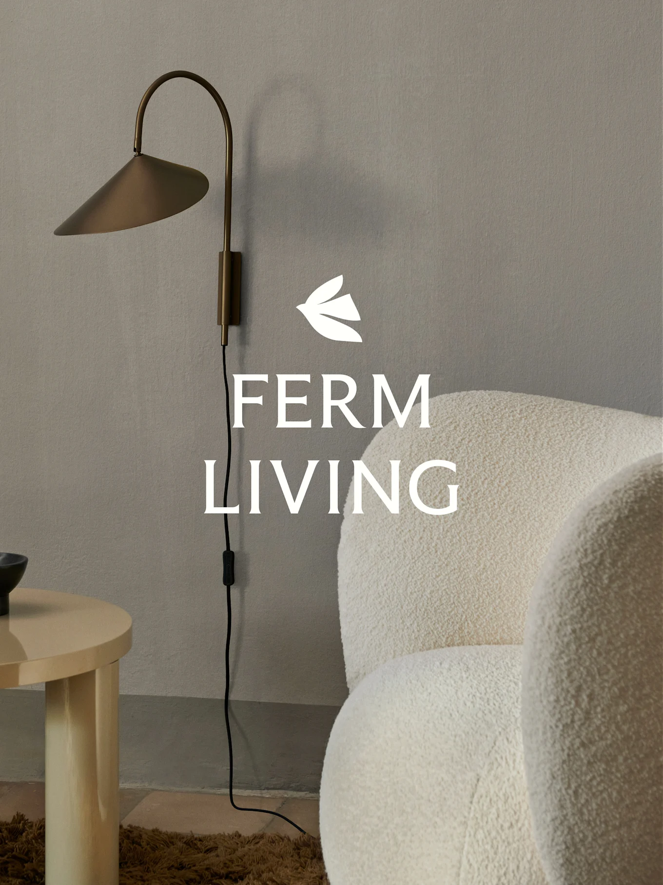 Ferm Living-logotyp över en vägglampa i mässing, en gräddfärgad bouclé-fåtölj och ett ljust sidobord i trä i en minimalistisk miljö.