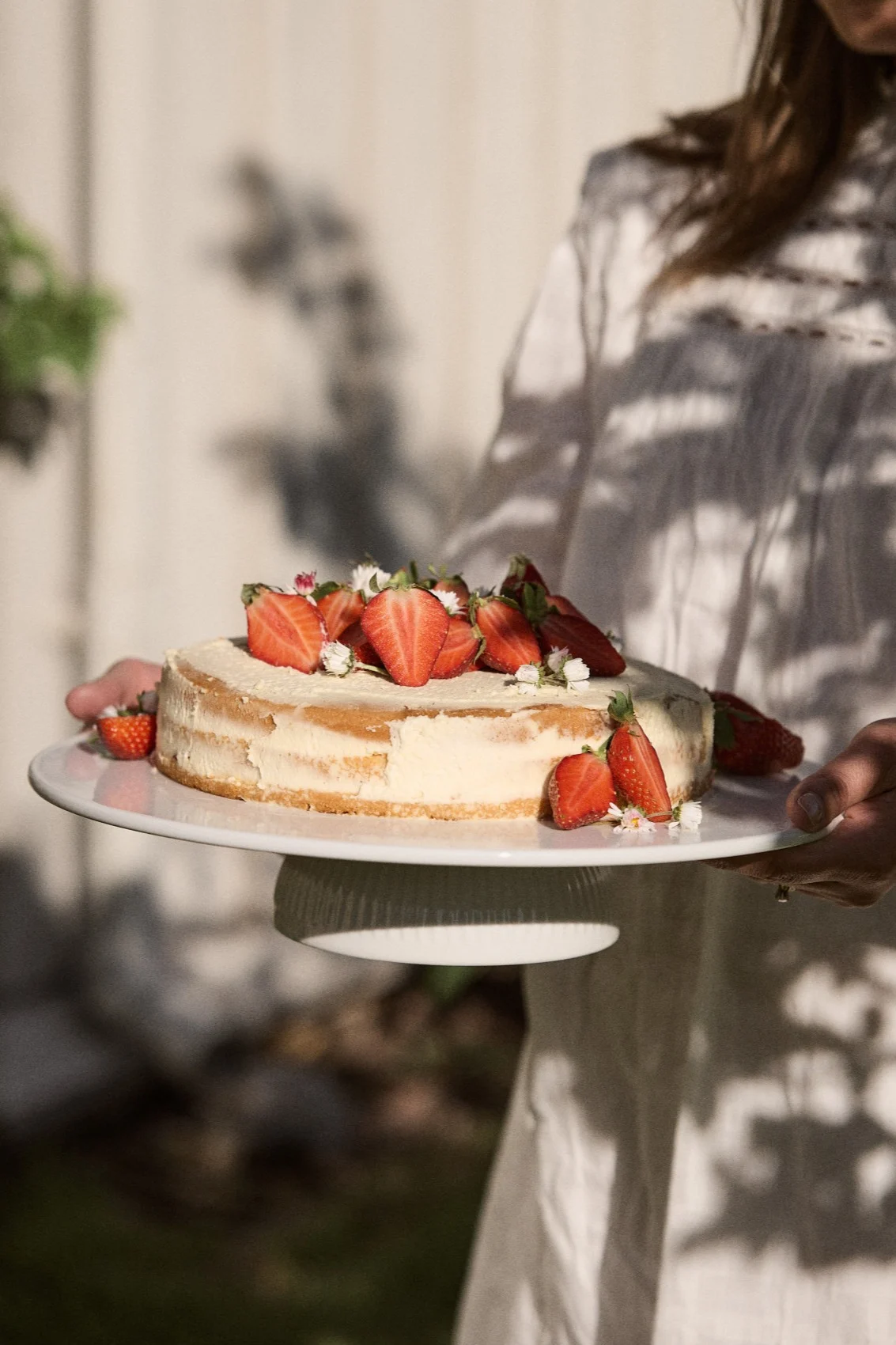 Person håller en rund tårta toppad med färska halverade jordgubbar och små vita blommor på ett vitt tårtfat utomhus.
