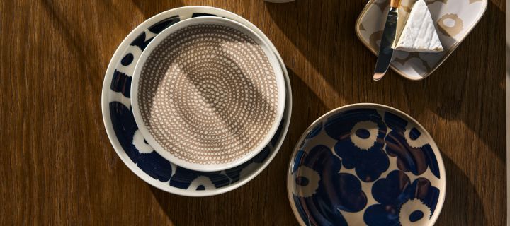 På ett träbord ligger Marimekkos Unikko tallrik i färgen vit-mörkblå, Unikko assiett i färgen terra-mörkblå, Unikko fat i beige och så en Siirtolapuutarha tallrik i färgen white-clay. 