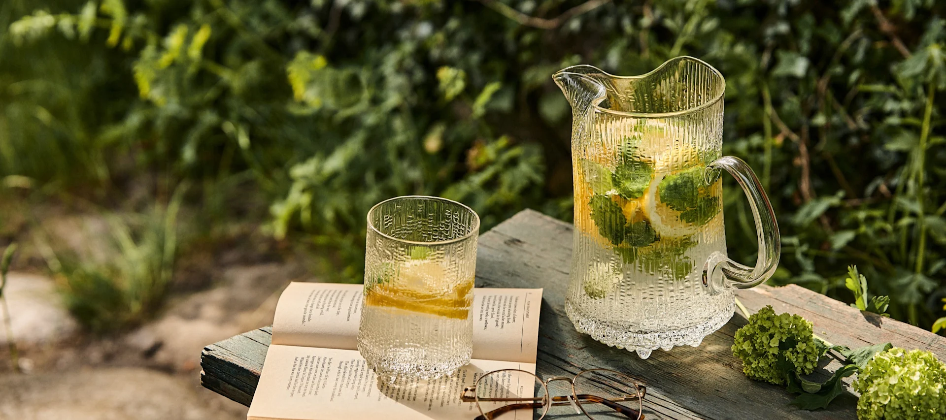 Texturerad glaskanna och glas med citron- och myntavatten, en öppen bok och glasögon på en träyta utomhus.