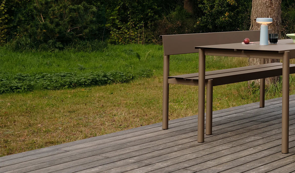 Modernt matbord och bänk i taupe utomhus på trädäck, med bärbar lampa och äpplen på bordet.