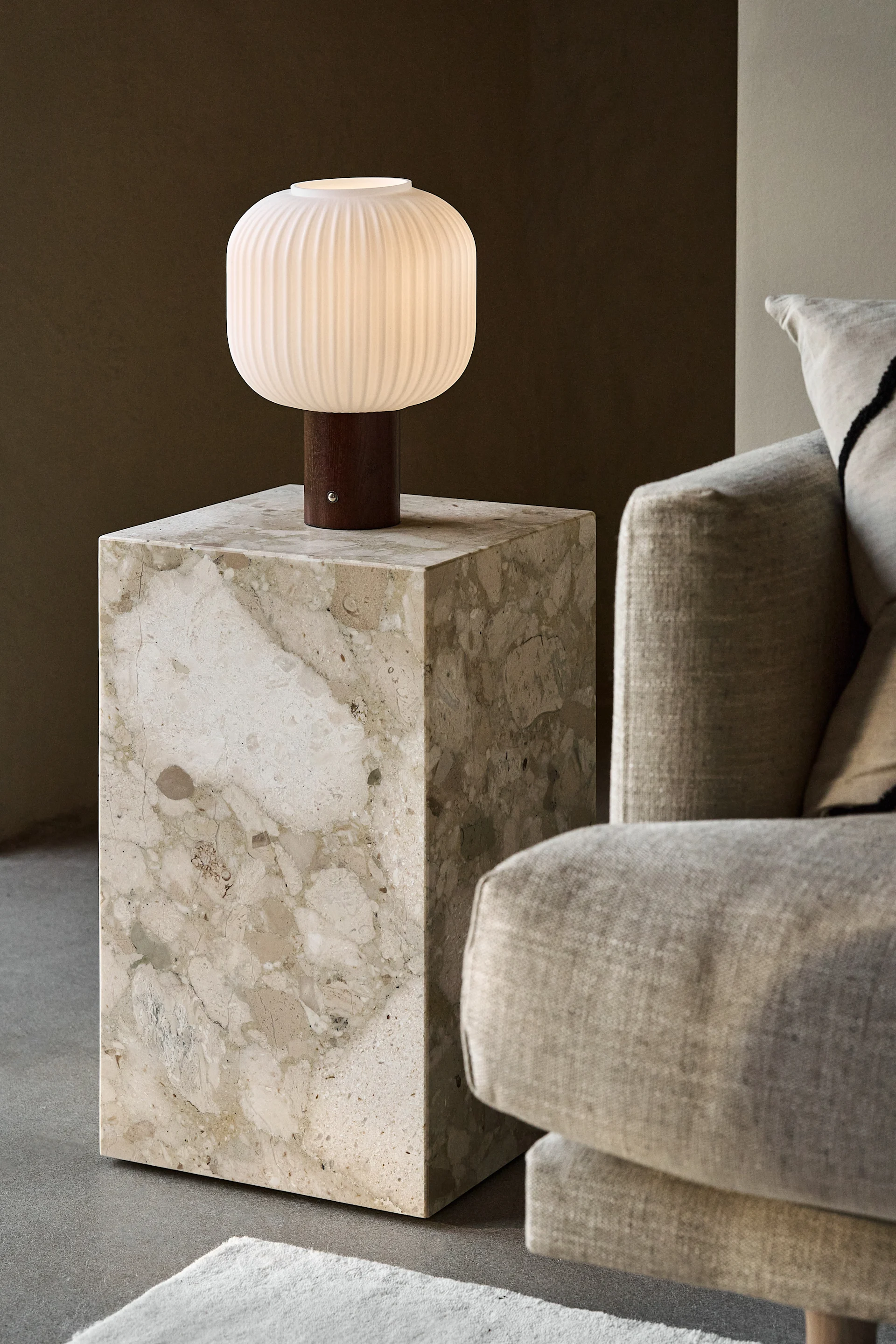 En tänd, räfflad, vit bordslampa med träfot på ett beige marmorbord bredvid en ljus soffa.