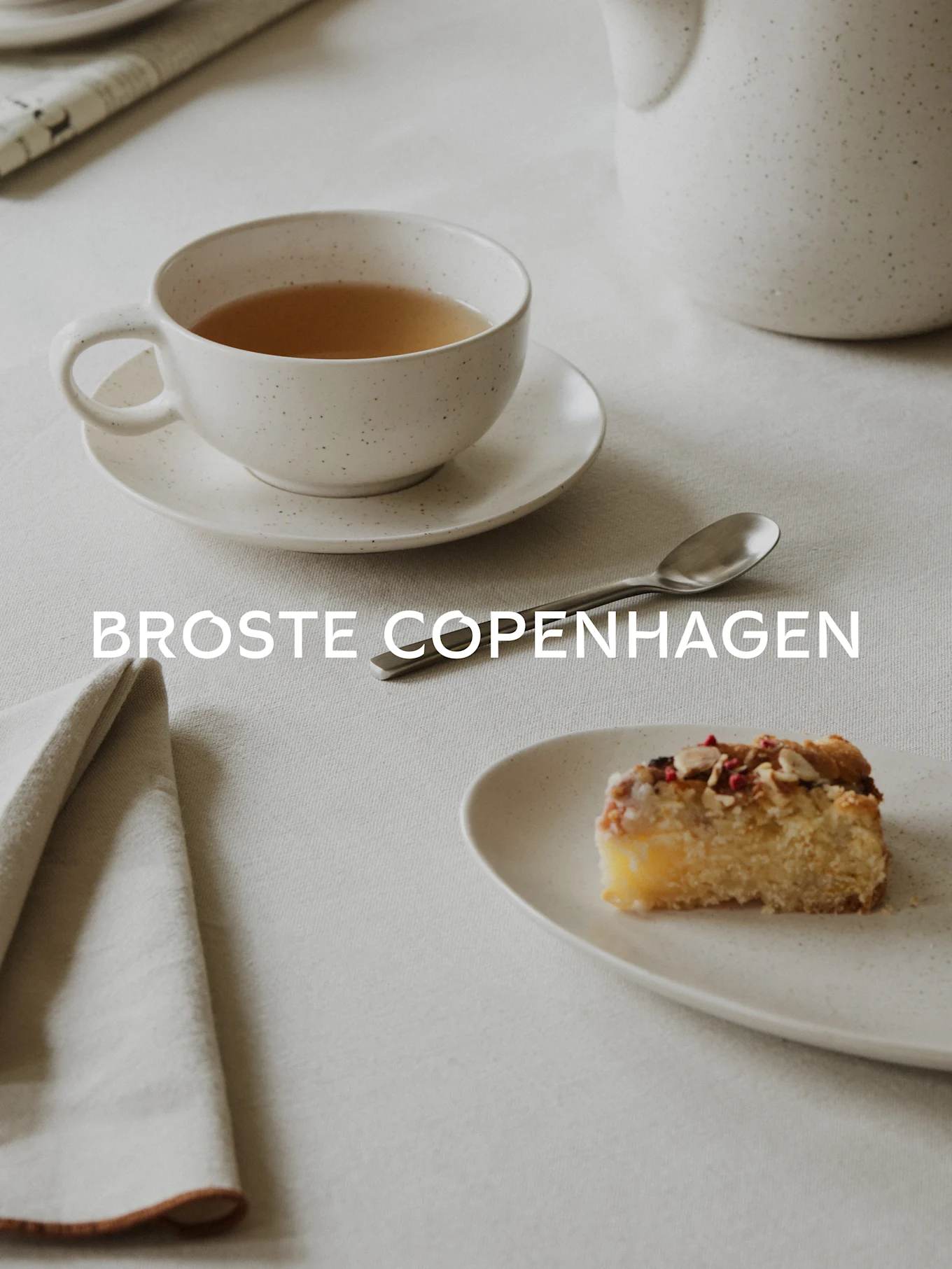 Te i en fläckig vit kopp, en bit kaka med mandlar och bär på en matchande tallrik, sked och servett på duk. Text: BROSTE COPENHAGEN.