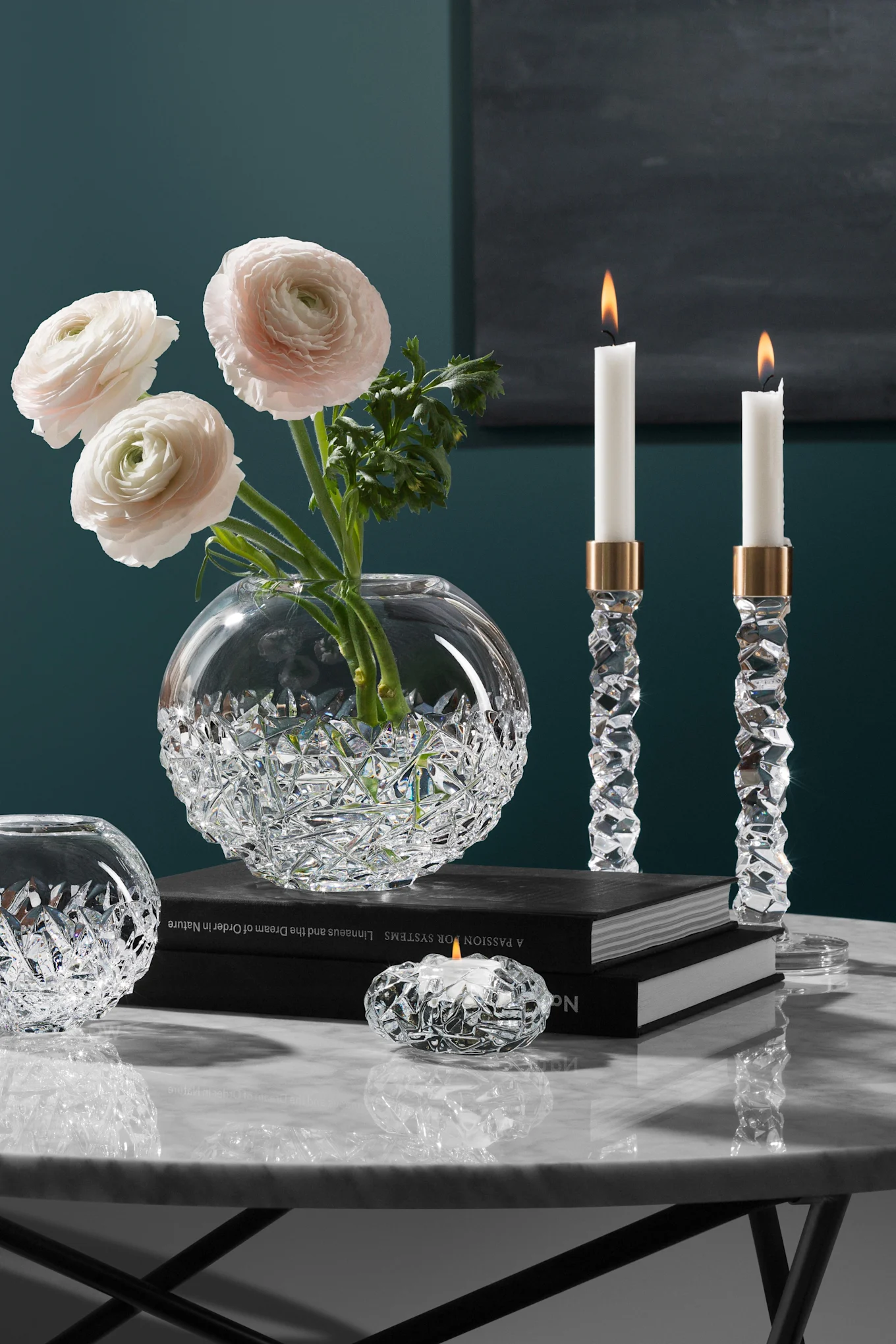 Elegant kristallvas med rosa ranunkel, två tända kristalljusstakar och en värmeljushållare på ett marmorbord.