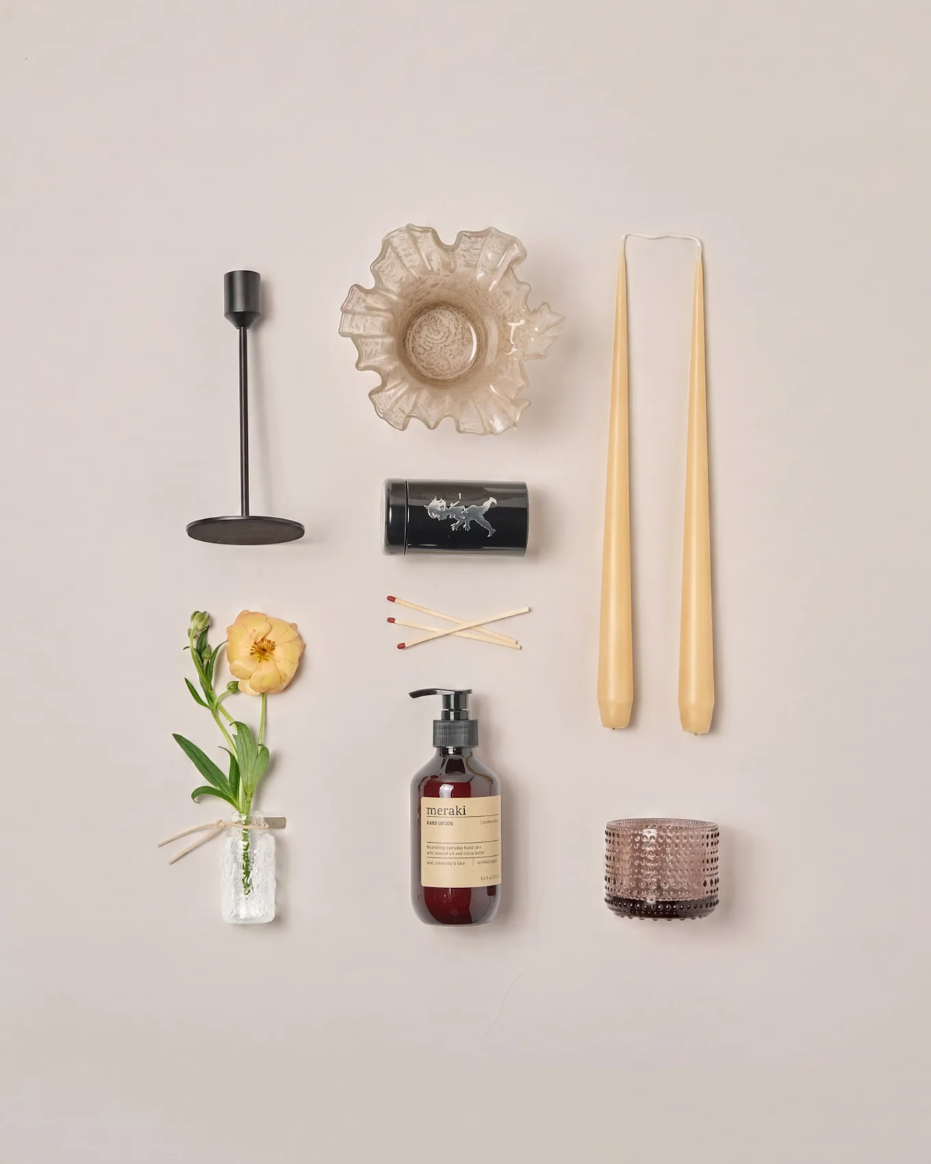 En platt lay av olika heminredningsartiklar: ljusstake, krusig skål, avsmalnande ljus, tändstickor, lotion, värmeljushållare och en ranunkelblomma.
