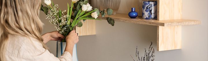 En kvinna står och justerar en blombukett som är nedstoppad i en Ada Stripe vas i vit färg med blåa organiskt formade linjer. Ovanför vasen på en hylla står också två blåa ljuslyktor. 