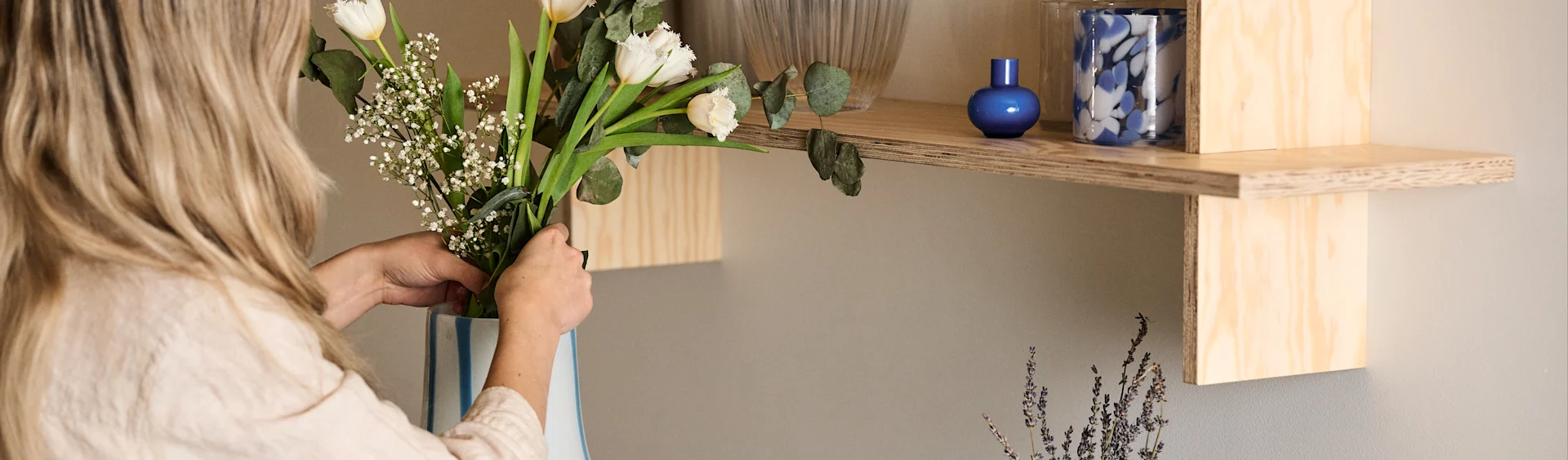 En kvinna står och justerar en blombukett som är nedstoppad i en Ada Stripe vas i vit färg med blåa organiskt formade linjer. Ovanför vasen på en hylla står också två blåa ljuslyktor.