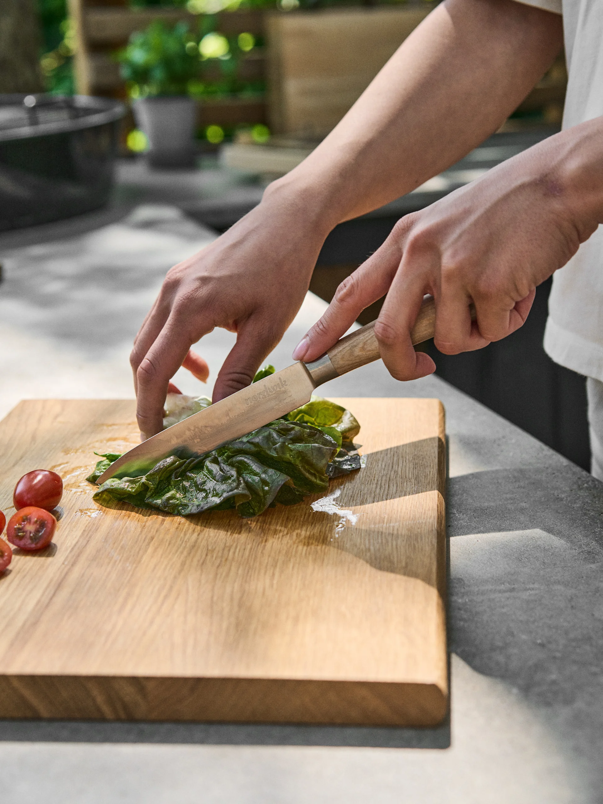 Händer skär ett grönt blad med en kniv på en träskärbräda, bredvid körsbärstomater.