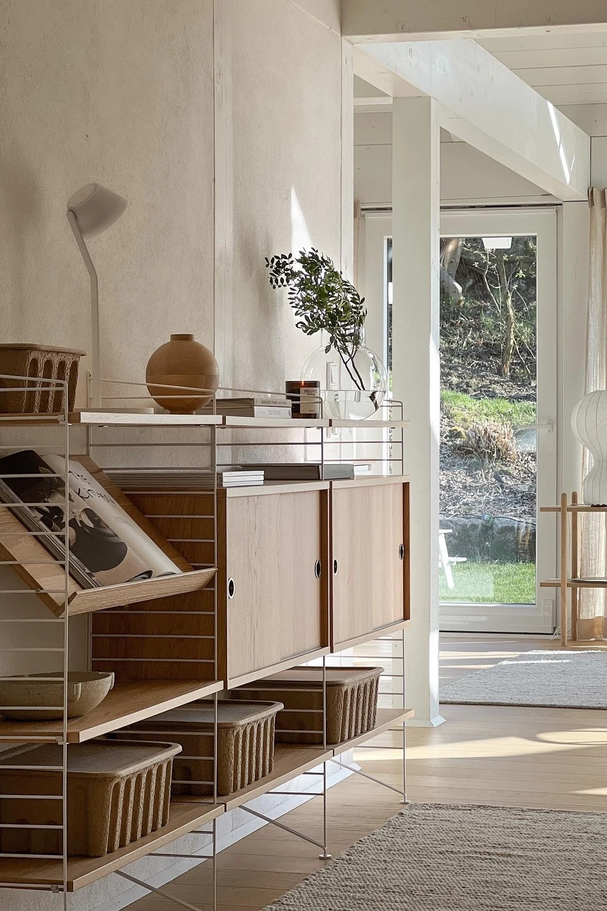 Ljust, modernt rum med en Stringhylla i ljust trä och vita ramar, fylld med vas, böcker, växter och korgar, med trädgårdsutsikt. Hyllan passar perfekt att dekorera vägg med. 