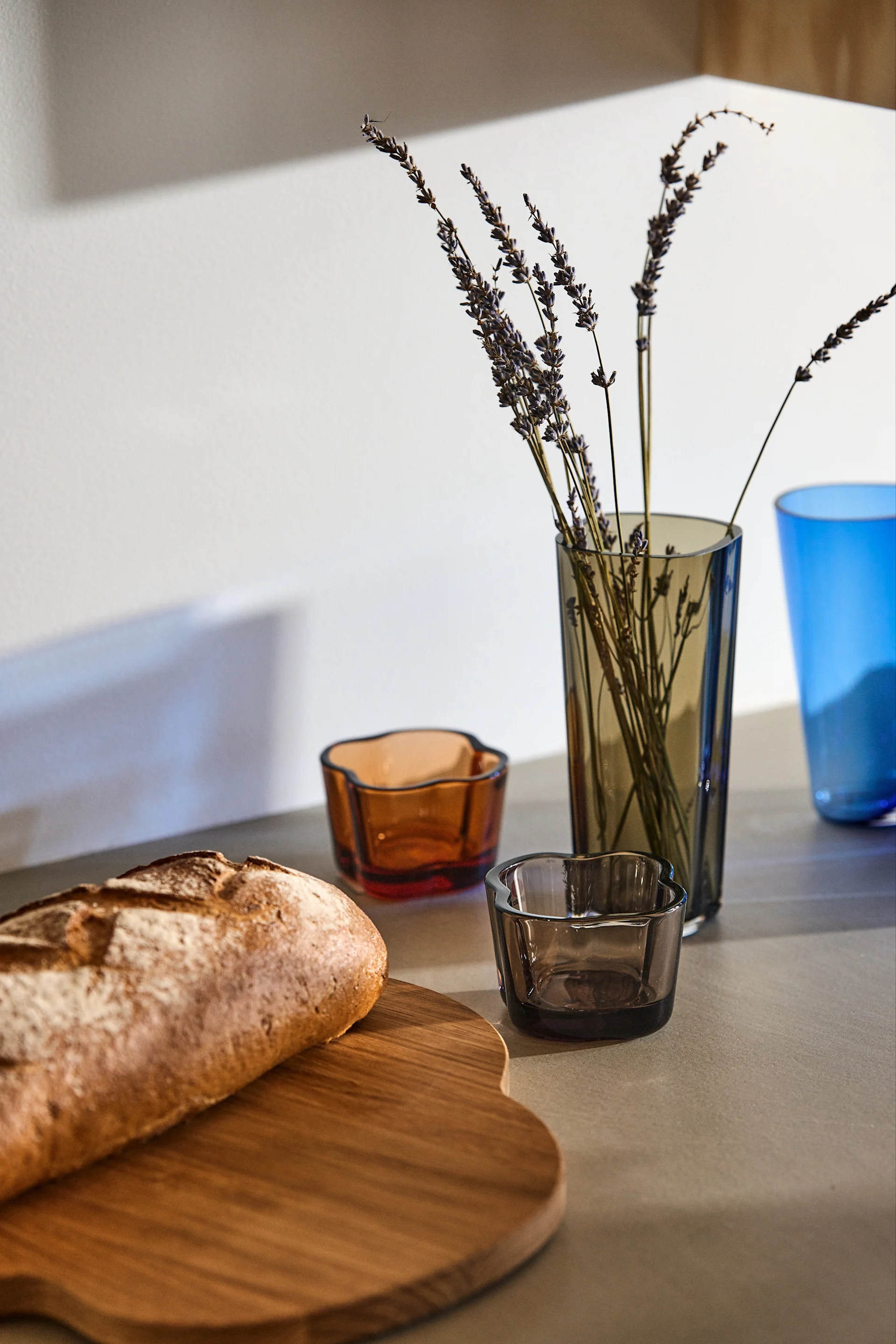 På en köksbänk bredvid en träbricka med en brödlimpa på, står två Alvar Aalto ljuslyktor från Iittala i färgerna pomerans och grå. Där står även Alvar Aalto vas i färgen rökgrå. 