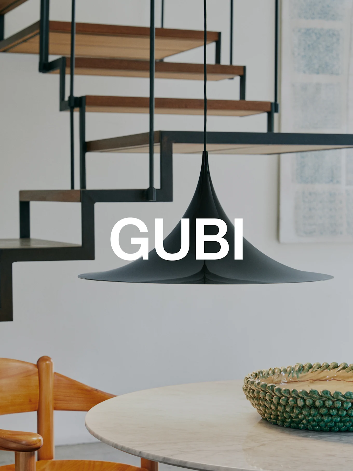 En svart konisk pendellampa över ett marmorbord med en grön skål och en trästol. En modern trappa i bakgrunden. Text 'GUBI'.