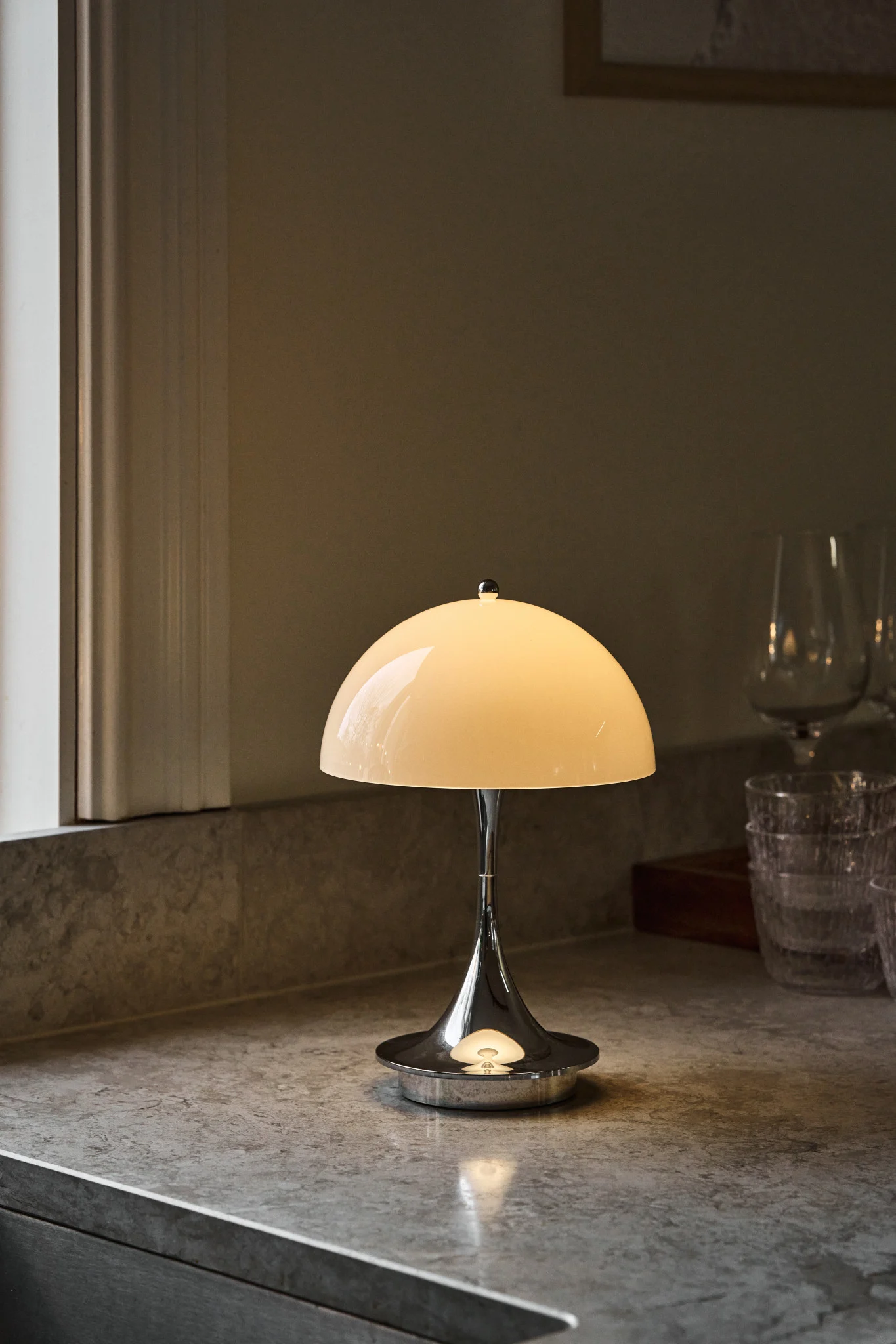Polerad krom bordslampa med glödande krämfärgad kupolskärm på en spräcklig grå bänkskiva, bredvid ett fönster och suddiga glas.